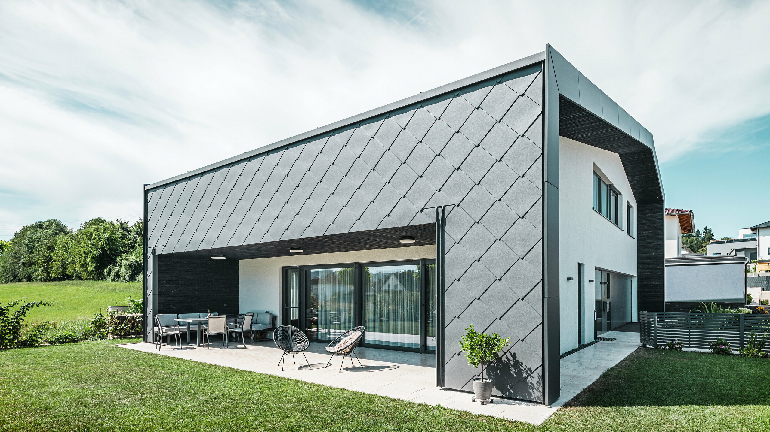 Välskött trädgård till en villa i Aschach. Byggnaden är stilfullt klädd med mörkgrå PREFA Väggromb 44 × 44 i aluminium. Den täckta terrassen erbjuder en mysig sittplats med utsikt över den grönskande gräsmattan. Den eleganta aluminiumklädseln i kombination med vita putsade ytor ger en modern och väderbeständig arkitektur. Stora fönsterpartier släpper in mycket dagsljus i rummen och skapar en öppen, ljus atmosfär.