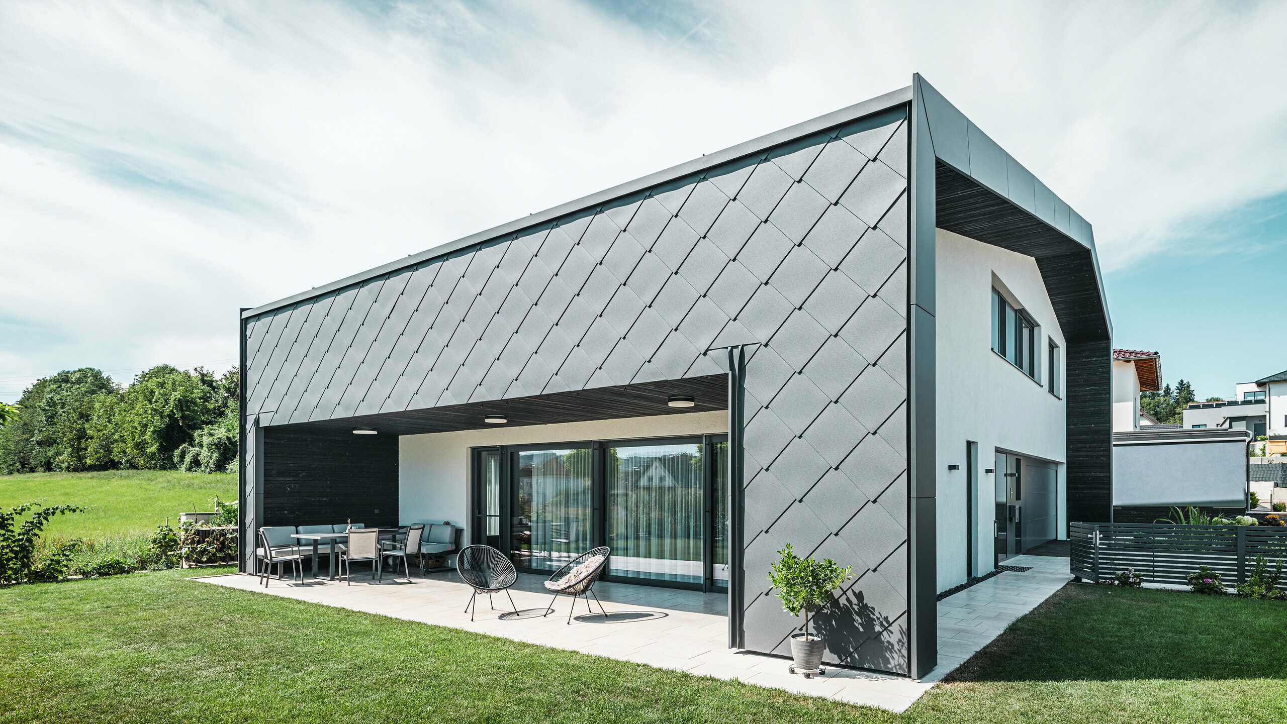 Välskött trädgård till en villa i Aschach. Byggnaden är stilfullt klädd med mörkgrå PREFA Väggromb 44 × 44 i aluminium. Den täckta terrassen erbjuder en mysig sittplats med utsikt över den grönskande gräsmattan. Den eleganta aluminiumklädseln i kombination med vita putsade ytor ger en modern och väderbeständig arkitektur. Stora fönsterpartier släpper in mycket dagsljus i rummen och skapar en öppen, ljus atmosfär.