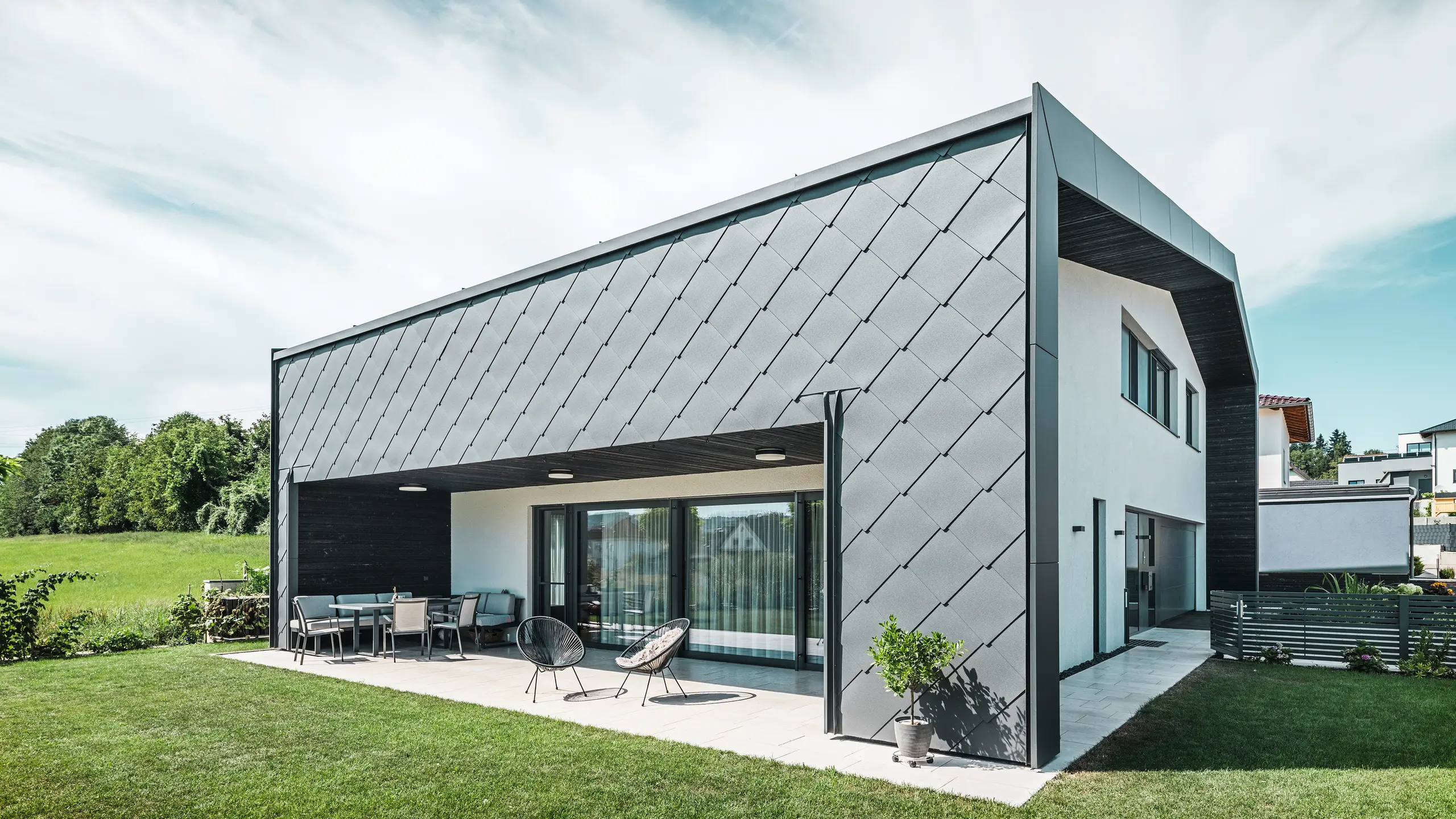Välskött trädgård till en villa i Aschach. Byggnaden är stilfullt klädd med mörkgrå PREFA Väggromb 44 × 44 i aluminium. Den täckta terrassen erbjuder en mysig sittplats med utsikt över den grönskande gräsmattan. Den eleganta aluminiumklädseln i kombination med vita putsade ytor ger en modern och väderbeständig arkitektur. Stora fönsterpartier släpper in mycket dagsljus i rummen och skapar en öppen, ljus atmosfär.