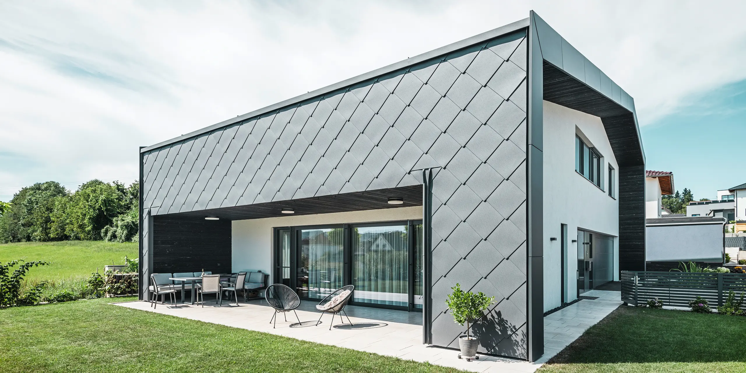 Välskött trädgård till en villa i Aschach. Byggnaden är stilfullt klädd med mörkgrå PREFA Väggromb 44 × 44 i aluminium. Den täckta terrassen erbjuder en mysig sittplats med utsikt över den grönskande gräsmattan. Den eleganta aluminiumklädseln i kombination med vita putsade ytor ger en modern och väderbeständig arkitektur. Stora fönsterpartier släpper in mycket dagsljus i rummen och skapar en öppen, ljus atmosfär.