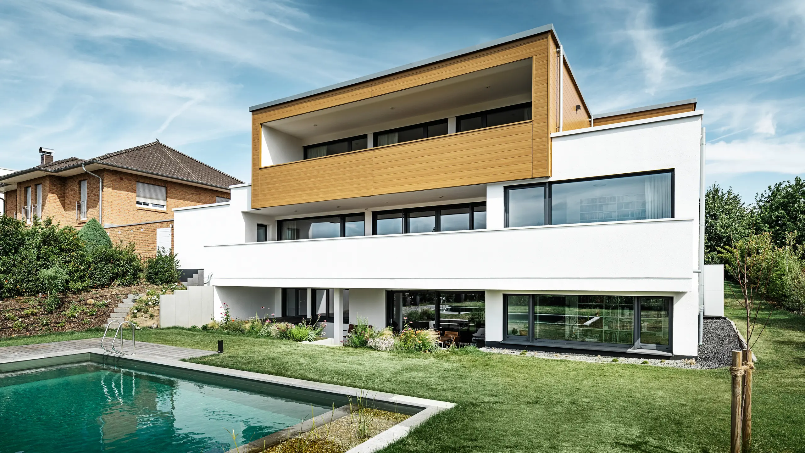 Baksidan av en modern villa i Usingen, Tyskland. Den gröna gräsmattan, en pool och ett ungt trädgårdsträd syns längst ner i bilden. Det iögonfallande huset har en vit byggnadsgrund av en putsad fasad, en aluminiumfasad på övervåningen av PREFA fasadpanel i ek natur och stora fönsterpartier med antracitfärgad ram. På avstånd syns grannhuset med tegelfasad i bildens vänstra kant.