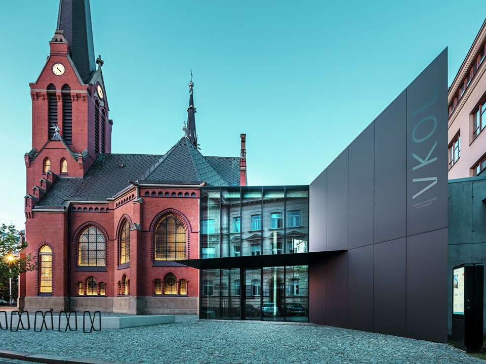 Das Bild zeigt das imposante Zusammenspiel zwischen der historischen Roten Kirche und dem modernen Neubau in Olmütz. Die Kirche mit ihren charakteristischen gotischen Fenstern und dem markanten Turm bildet das historische Rückgrat des Ensembles, während der moderne Anbau mit seiner eleganten, schwarzen PREFABOND Aluminium-Verbundplatten-Fassade eine zeitgemäße, architektonische Antwort darstellt. Große Glasflächen lassen den Blick in das Innere des Neubaus zu, wo die Umgebung durch Reflexionen in den Fenstern eine interessante Wechselwirkung zwischen Alt und Neu widerspiegelt. Die präzise Verarbeitung der PREFABOND-Platten betont die moderne Ästhetik, während die harmonische Integration in das historische Umfeld sowohl funktional als auch visuell beeindruckt.