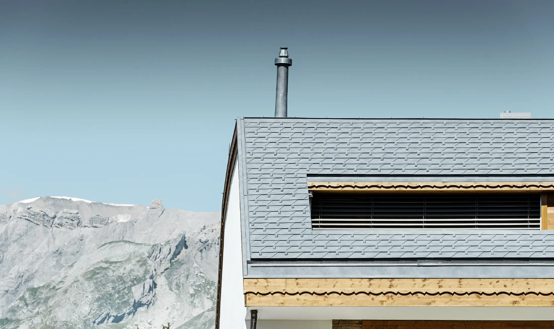 Flerfamiljshus i Crans-Montana med utsikt över bergen och en fasad med lekfulla träelement med ett tak av PREFA:s takshingel i stengrå aluminium