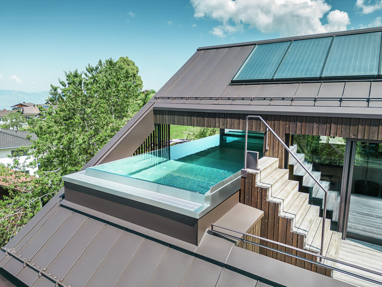 Luftaufnahme eines modernen Hauses mit einem nussbraunen PREFA Prefalz Dach. Eine Dachterrasse mit Pool bietet einen exklusiven Entspannungsbereich mit Blick auf die hügelige, grüne Landschaft.