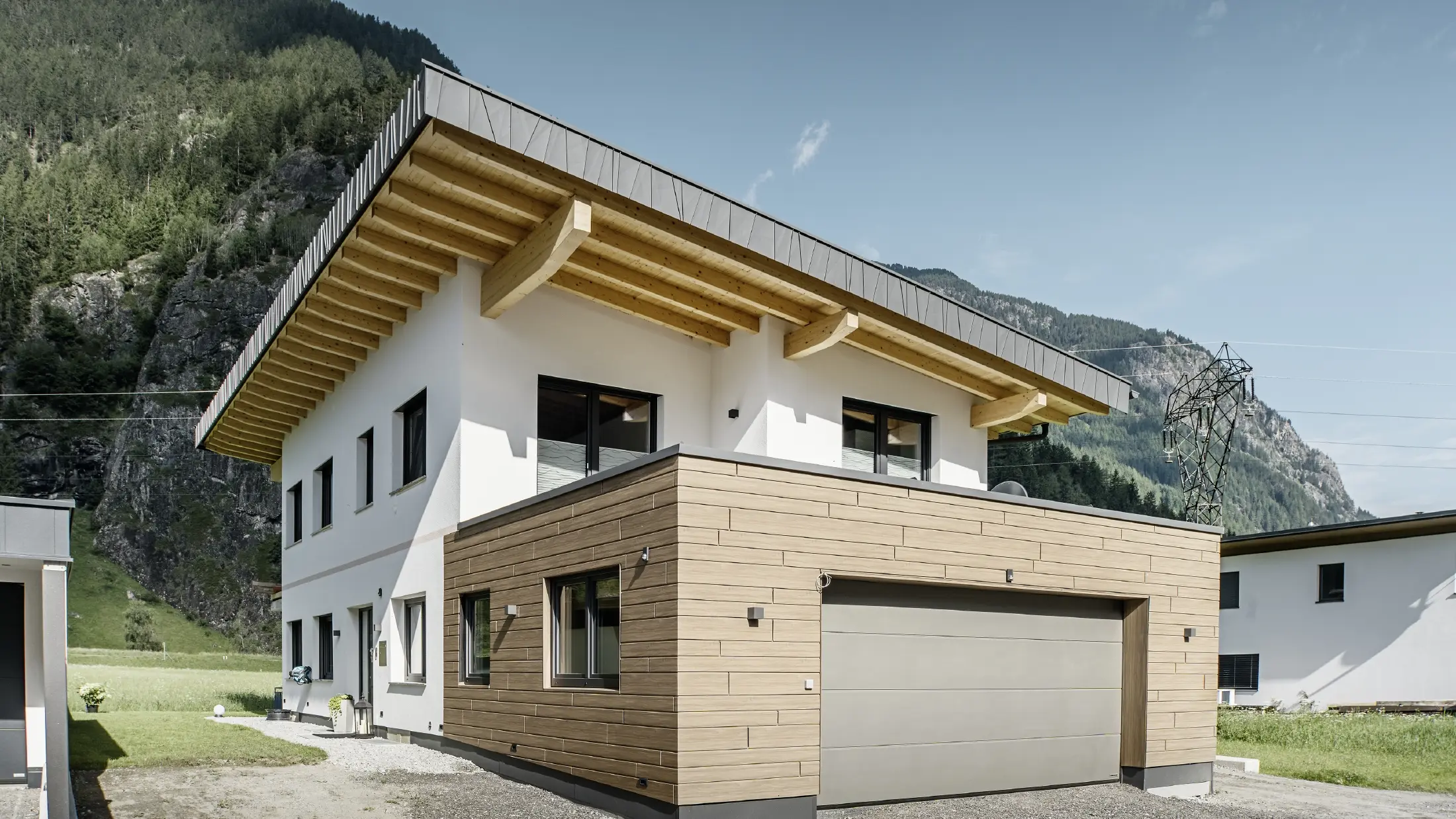 Modernt enfamiljshus med renoverad fasad: nyrenoverad ytterfasad med horisontell fasadbeklädnad i träimitation, kombinerad med vitputsade ytor, integrerad garagebeklädnad och modern arkitektur i alpin miljö.