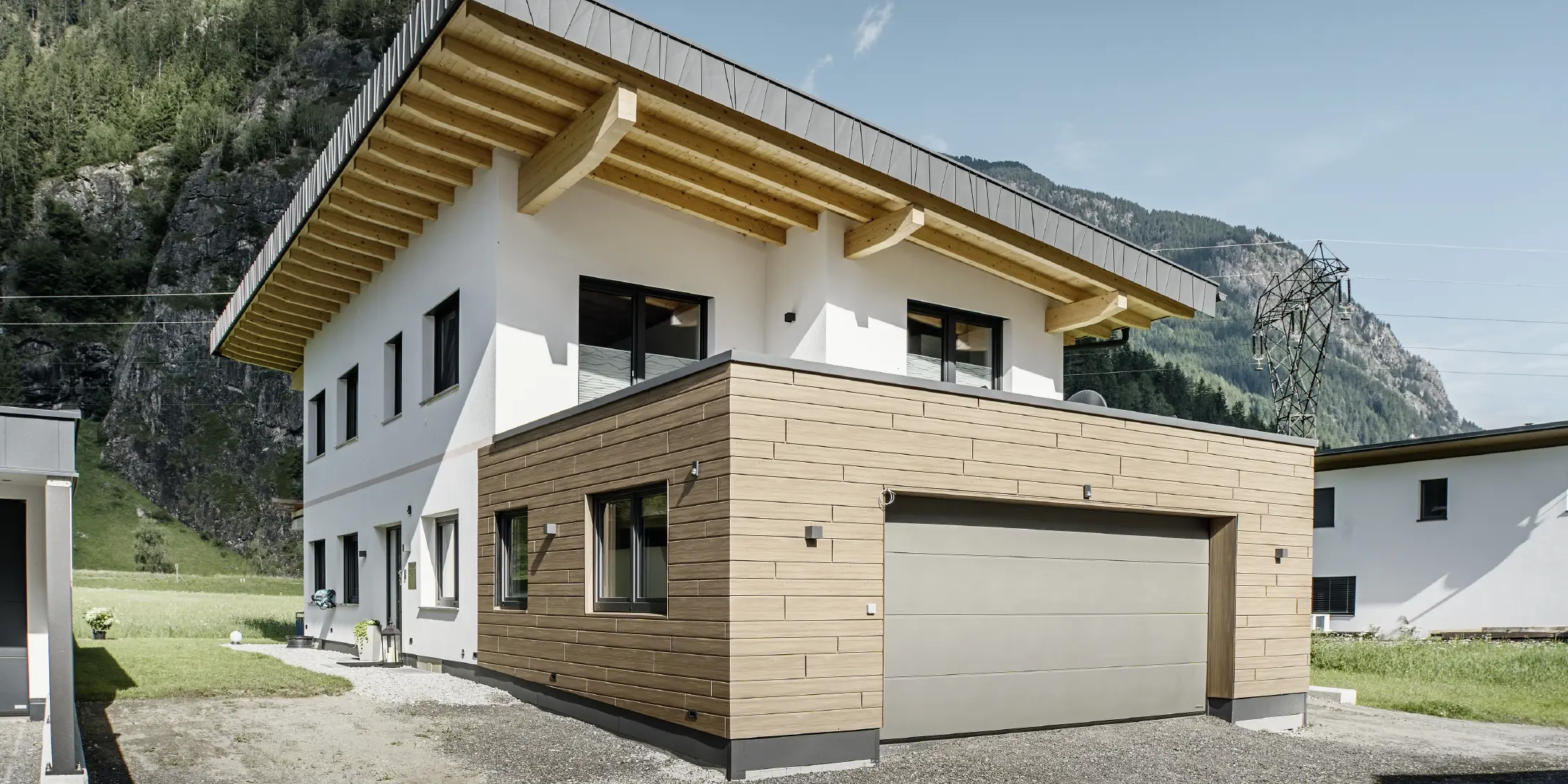 Modernt enfamiljshus med renoverad fasad: nyrenoverad ytterfasad med horisontell fasadbeklädnad i träimitation, kombinerad med vitputsade ytor, integrerad garagebeklädnad och modern arkitektur i alpin miljö.