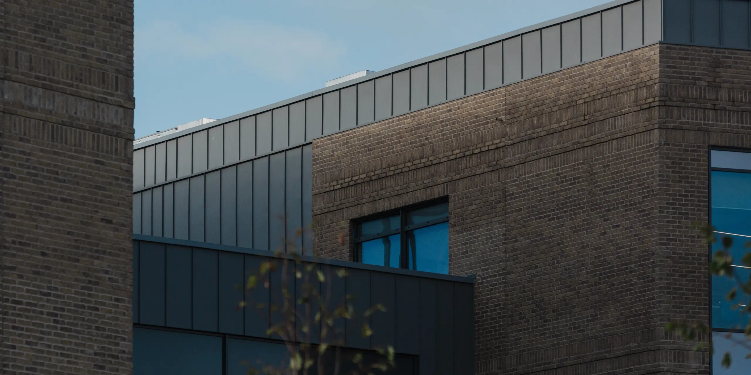 Ein präziser Fassadendetailblick auf das Northern Regional College in Ballymena zeigt den oberen Gebäudeabschluss mit vertikal verlegten PREFALZ Aluminium-Stehfalzbahnen in Patinagrau. Die klaren Linien des Aluminiumbandes kontrastieren wirkungsvoll mit dem warmen Klinkermauerwerk und den tief eingeschnittenen Fensteröffnungen. Die ruhige Metalloberfläche reflektiert das Tageslicht subtil vor blauem Himmel. PREFA Aluminium überzeugt hier durch Langlebigkeit, formale Präzision und eine zeitlose, wartungsarme Lösung für moderne Bildungsbauten.