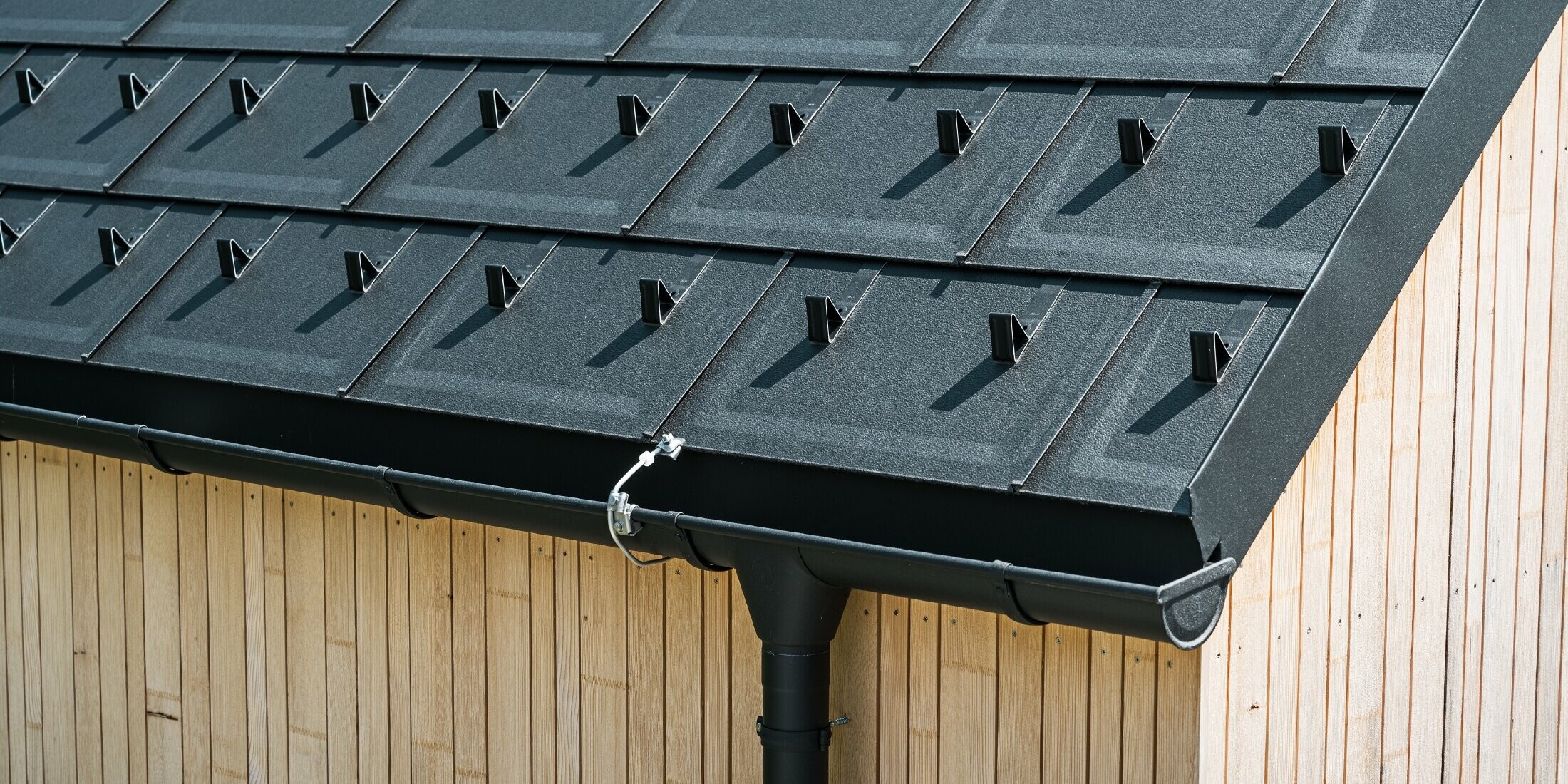 Närbild på PREFA takplatta R.16 med snöstopp, takränna och stuprör i P.10 svart inkl. åskskydd och en lodrät träfasad