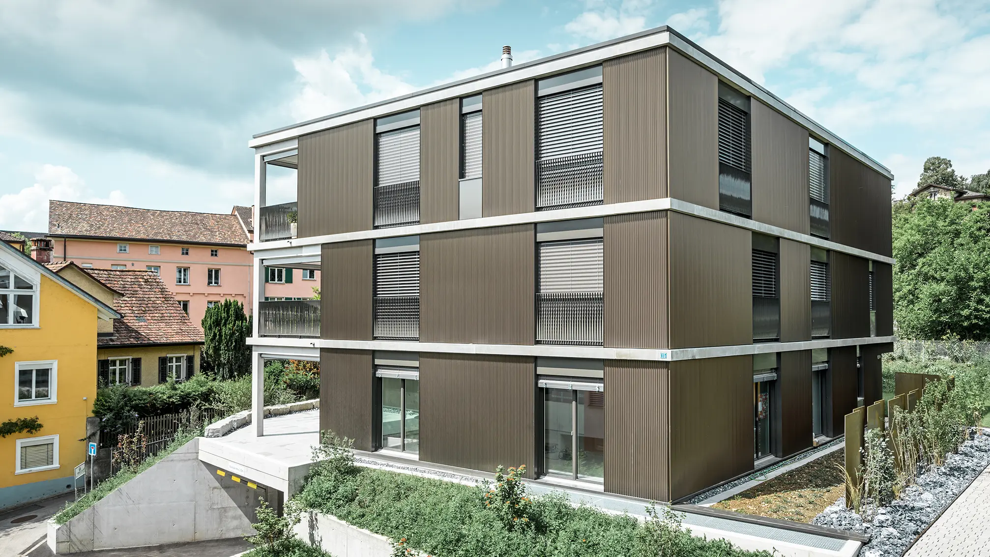 Den markanta aluminiumfasaden på detta flerfamiljshus i Schaffhausen är utrustad med det eleganta PREFA-tandprofilen i färgen Colinal. Den vertikala placeringen av de strängpressade aluminiumprofilerna gör att byggnaden ser mer kompakt ut och fasadstrukturen får en harmonisk balans. Den eleganta designen smälter smakfullt in i omgivningen och ger byggnaden en modern elegans som är både visuellt tilltalande och funktionell. Perspektivet visar flerfamiljshuset från framsidan med garageinfarten. I bakgrunden syns träd, andra byggnader och en kyrktorn.