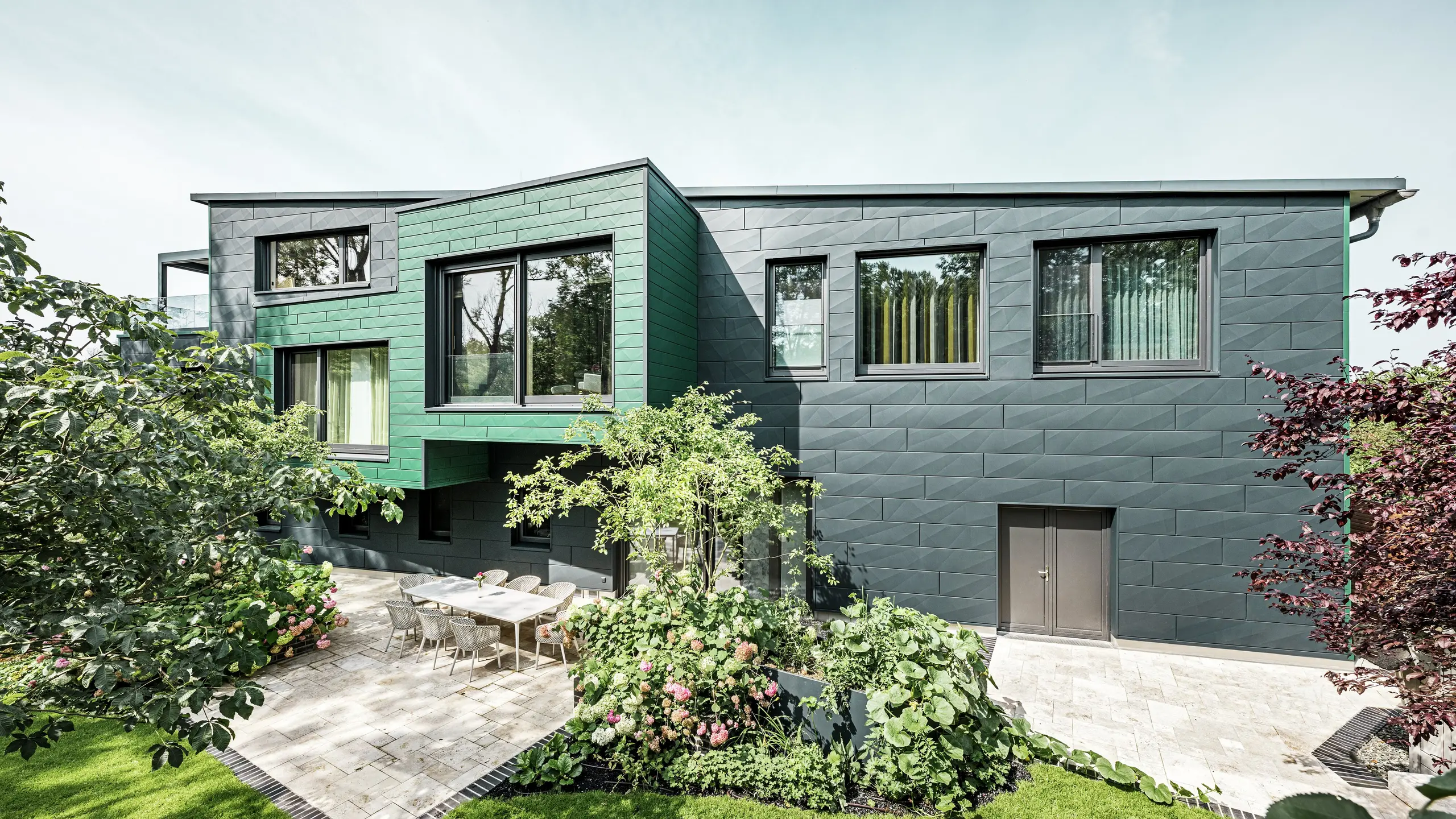 Enfamiljshuset i Türkheim imponerar med sin extraordinära fasad, som är klädd med PREFA Siding X-paneler i färgerna antracit och mossgrön. De rena linjerna och det markanta färgvalet på aluminiumpanelerna ger byggnaden ett modernt och elegant utseende. I förgrunden syns den generösa trädgården med en stilfull terrass och ett matbord med moderna stolar. Färgglada rabatter och friska växter skapar en livfull, grön kontrast till den robusta fasaden. Den hållbara och underhållsvänliga aluminiumfasaden skyddar byggnaden och ger den en tidlös skönhet.