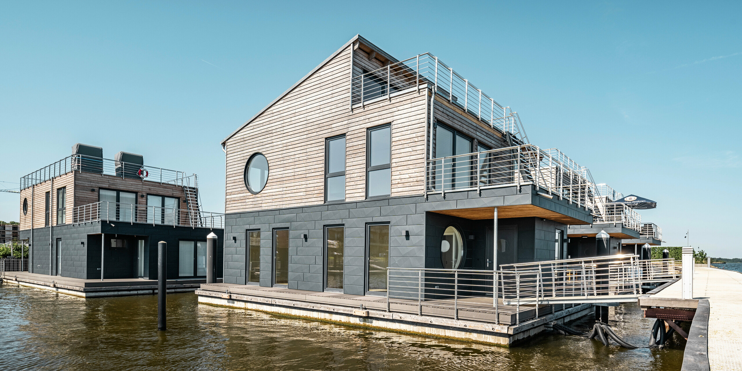 Ein architektonisches Ensemble von Wasserhäusern im Pionierhafen Schleswig, charakterisiert durch die Kombination von PREFA Siding.X Aluminiumfassaden in P.10 Anthrazit und natürlichen Holzelementen. Die Gebäude mit ihren großen runden und rechteckigen Fenstern ermöglichen einen Panoramablick auf die Schlei und sind durch Stege zugänglich, die eine nahtlose Verbindung zum Festland schaffen. Die Dachterrassen und die direkte Lage am Wasser reflektieren eine luxuriöse und maritime Lebensweise.
