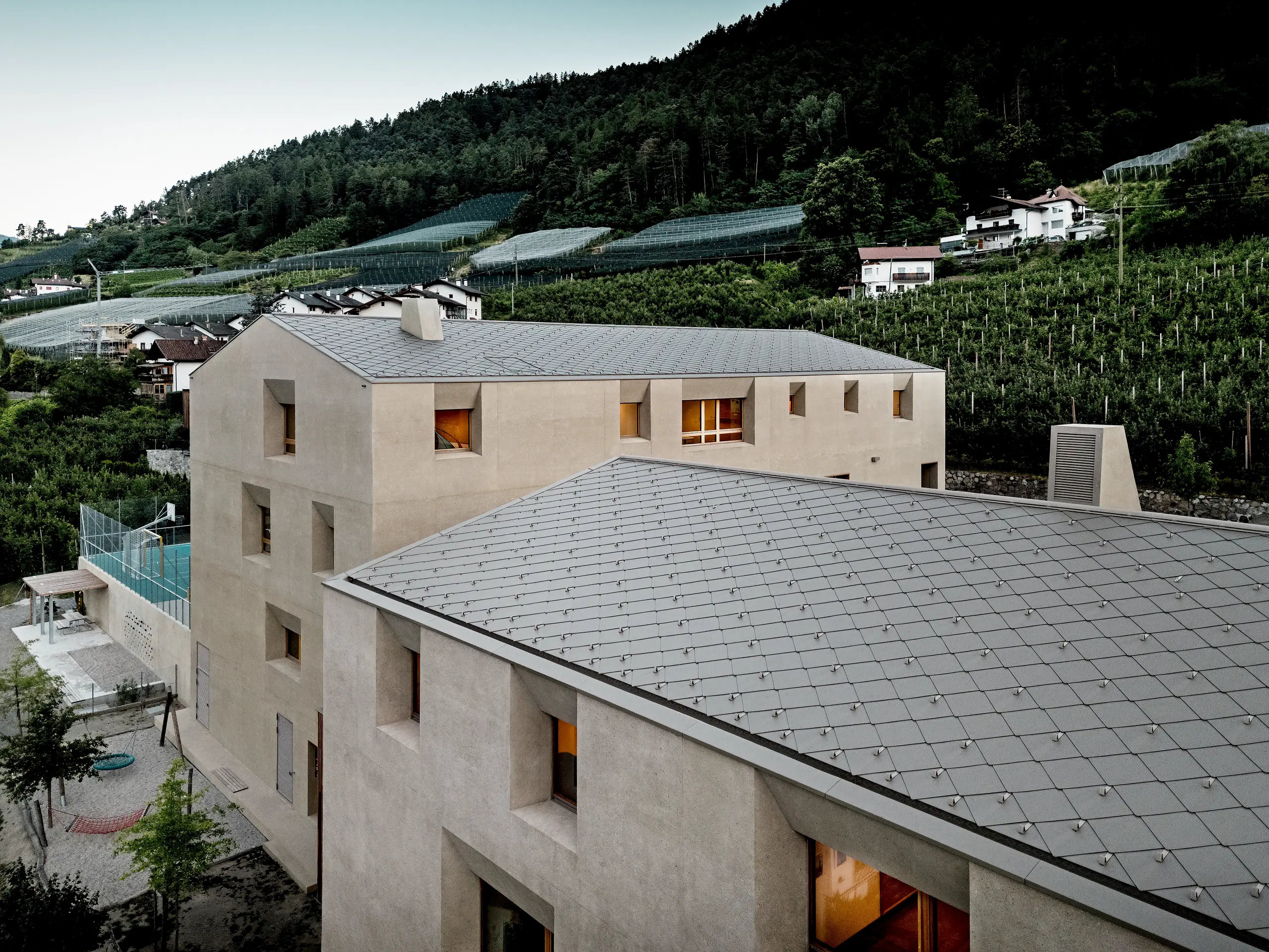 Aus der Vogelperspektive zeigt das Bildungszentrum Riffian in Südtirol seine markanten PREFA Dachrauten 44 × 44 in P.10 Bronze. Die präzise Aluminiumdeckung spannt sich ruhig über Kindergarten und Volksschule, schützt wetterfest und langlebig und fügt die moderne Architektur eindrucksvoll in Obstgärten und Berghang ein.