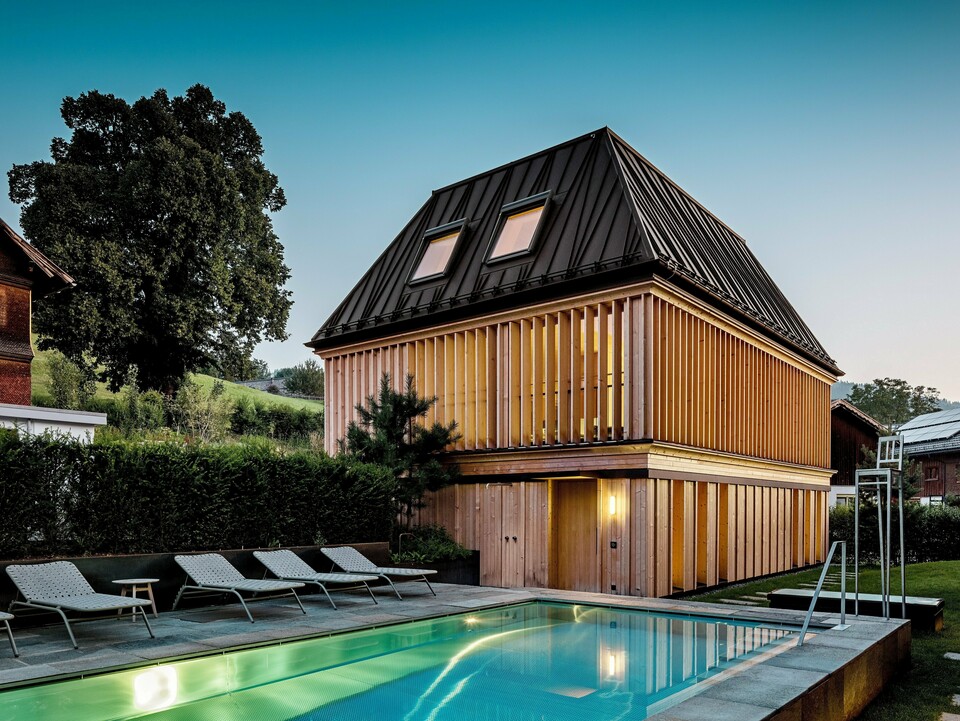 Das „Badehaus“ des Hotel Hirschen in Schwarzenberg in der Dämmerung, mit Blick auf den beleuchteten Außenpool im Vordergrund. Das Gebäude beeindruckt durch sein modernes Walmdach aus PREFALZ in der Farbe P.10 Nussbraun mit präzise ausgeführten, teils schräg verlaufenden Stehfalzen. Zwei Dachfenster sorgen für Tageslicht im Inneren. Die Fassade besteht aus vertikalen, geschoßhohen Holzlamellen, die dem Baukörper Struktur und Leichtigkeit verleihen. Im unteren Bereich dominiert naturbelassenes Holz. Umgeben von gepflegtem Grün und eingebettet in die Hügellandschaft des Bregenzerwaldes entsteht eine harmonische Verbindung aus Architektur, Natur und Erholung.
