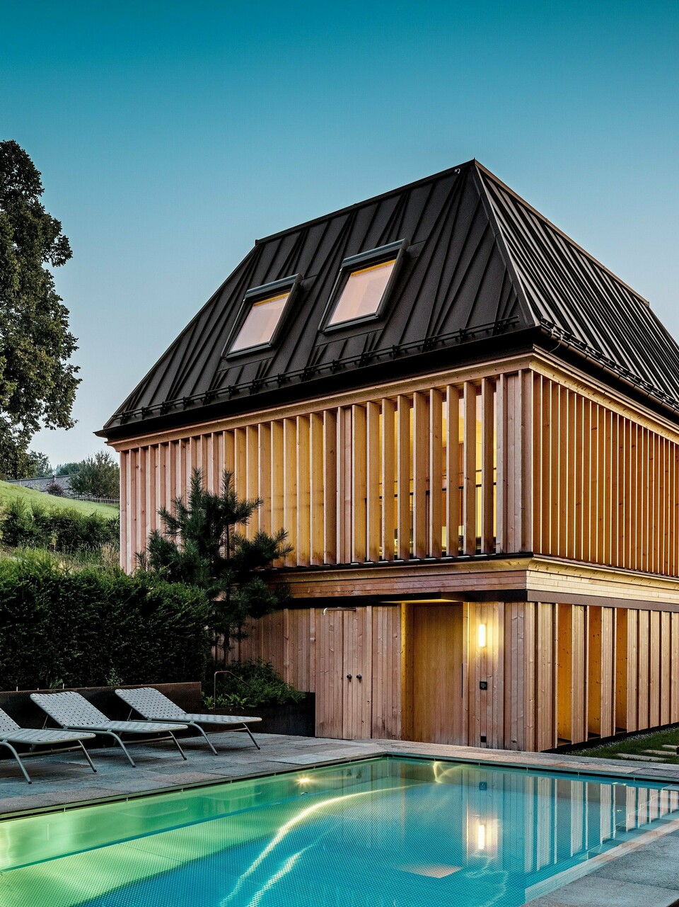 Das „Badehaus“ des Hotel Hirschen in Schwarzenberg in der Dämmerung, mit Blick auf den beleuchteten Außenpool im Vordergrund. Das Gebäude beeindruckt durch sein modernes Walmdach aus PREFALZ in der Farbe P.10 Nussbraun mit präzise ausgeführten, teils schräg verlaufenden Stehfalzen. Zwei Dachfenster sorgen für Tageslicht im Inneren. Die Fassade besteht aus vertikalen, geschoßhohen Holzlamellen, die dem Baukörper Struktur und Leichtigkeit verleihen. Im unteren Bereich dominiert naturbelassenes Holz. Umgeben von gepflegtem Grün und eingebettet in die Hügellandschaft des Bregenzerwaldes entsteht eine harmonische Verbindung aus Architektur, Natur und Erholung.