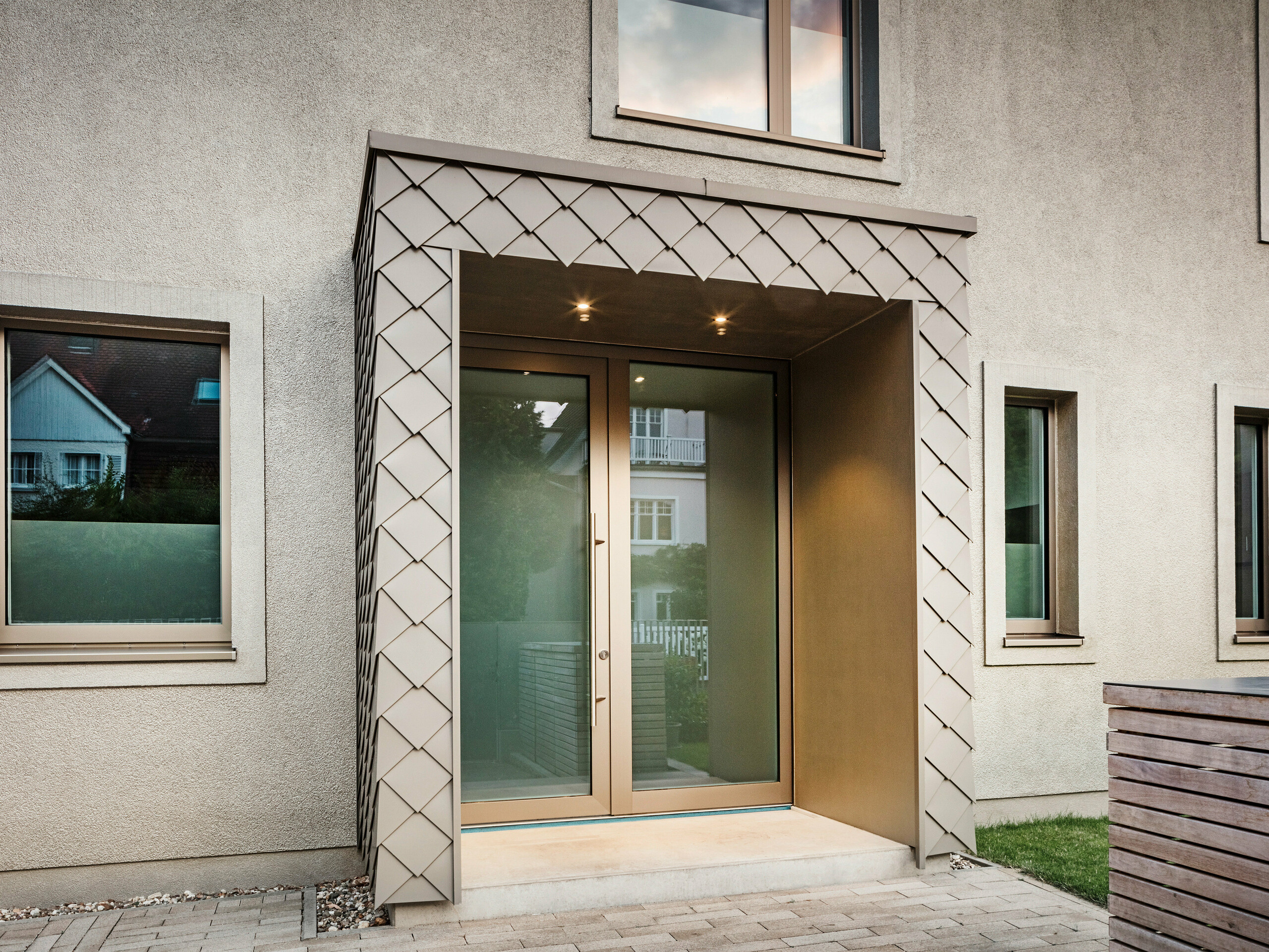 Detailaufnahme des Eingangsbereichs eines modernen Einfamilienhauses mit bronze-farbener Aluminium-Verkleidung von PREFA. Der Eingang ist überdacht und mit eleganten Aluminium-Rauten verkleidet, die dem Bereich einen modernen und stilvollen Look verleihen. Links und rechts neben der Eingangstür befinden sich große Fenster, die für eine helle und einladende Atmosphäre sorgen.