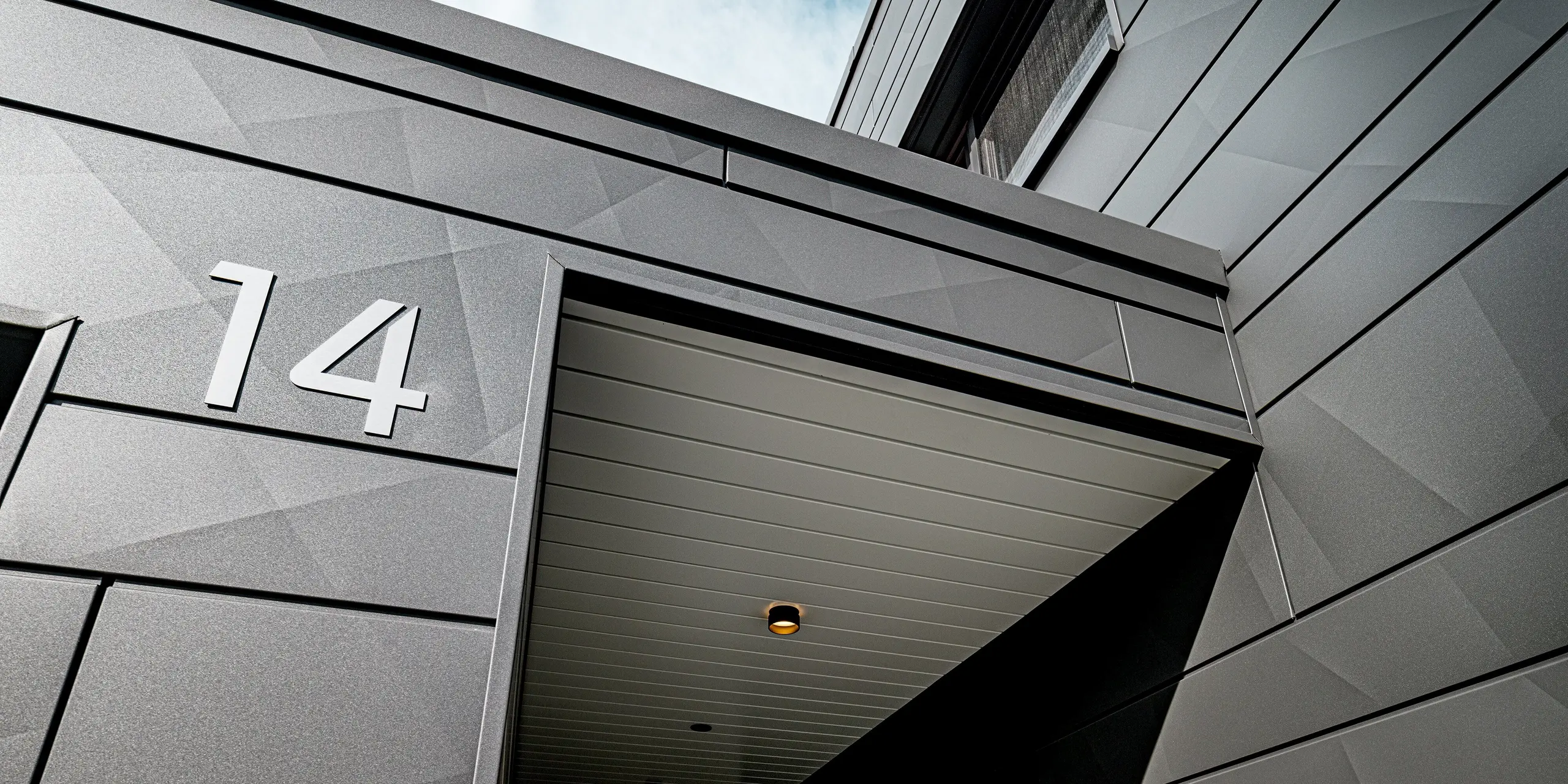 Detailansicht des überdachten Eingangs eines modernen Einfamilienhauses in Heidenheim mit PREFA Siding.X Fassade in P.10 Dunkelgrau. Die unterschiedlich gekanteten Aluminiumpaneele erzeugen ein markantes geometrisches Muster, das im Licht ein lebendiges Spiel aus Schatten und Reflexionen bildet. Die klar ausgeführte Hausnummer „14“ hebt sich präzise von der robusten Oberfläche ab. Die schützende Überdachung mit eingelassenen Spots unterstreicht die hochwertige Verarbeitung und zeigt die langlebige, witterungsbeständige Qualität der PREFA Aluminiumlösung.