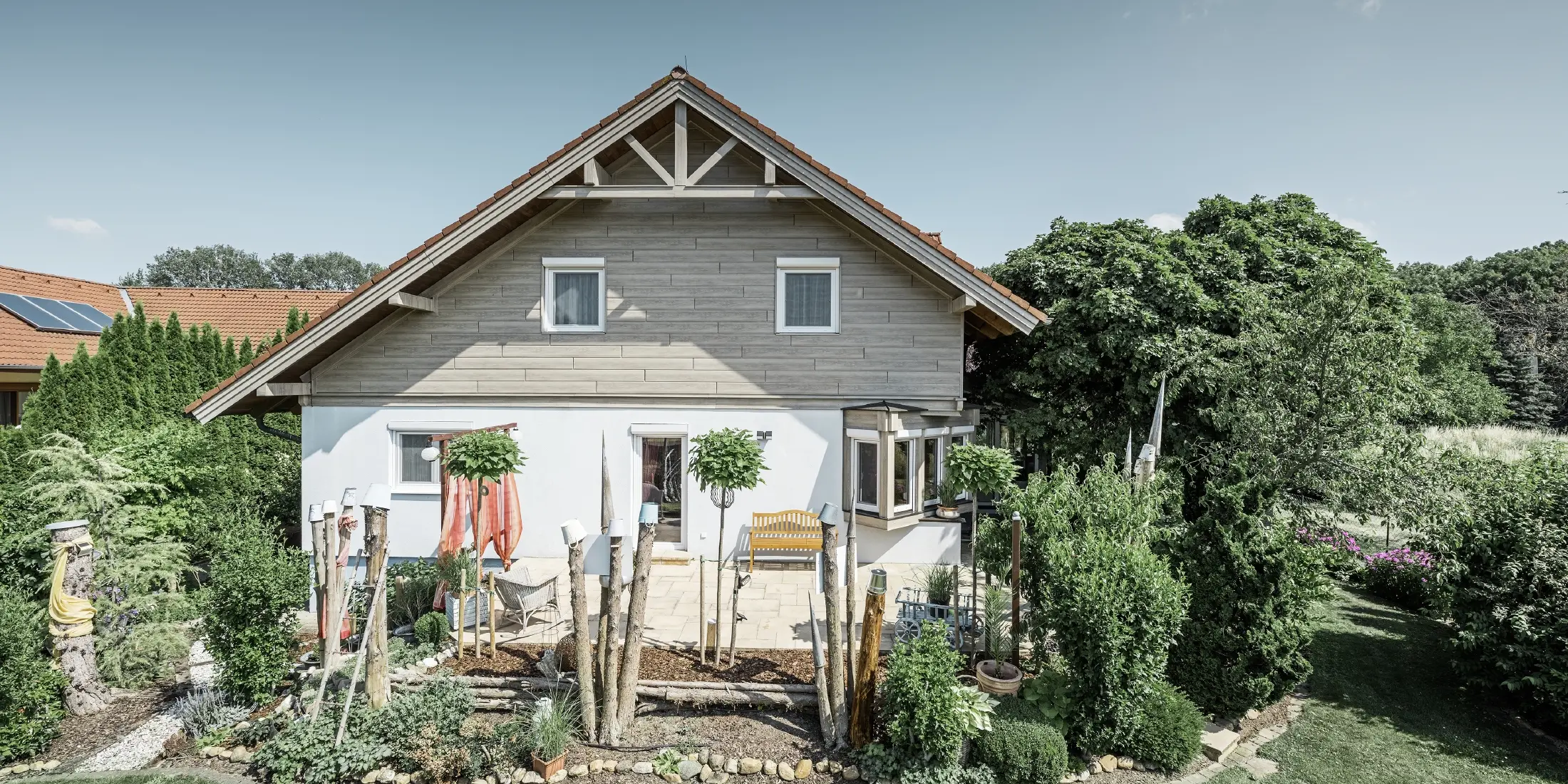 En villa med renoverad fasad och ny gavelbeklädnad i modern trälook i gråbeige, kombinerat med putsad fasad, sadeltak och en välskött trädgård.