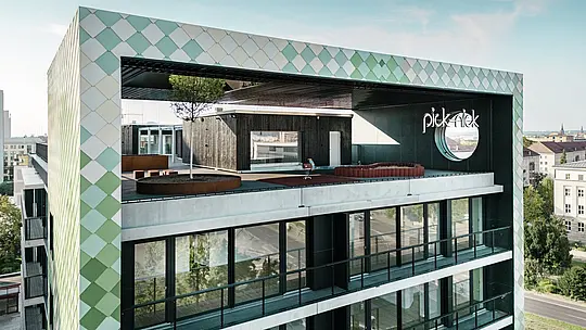 Detailansicht der Fassade des Wohn- und Geschäftshauses „Pick-Nick“ in Dresden mit seiner markanten PREFA-Aluminium-Rhombusfassade. Hauptattraktion ist die großzügige Dachterrasse mit Grünpflanzen, Sitzgelegenheiten und Kinderspielbereichen. Der Schriftzug „Pick-Nick“ und die innovative Fassadengestaltung setzen markante Akzente und verbinden Funktionalität mit Ästhetik.