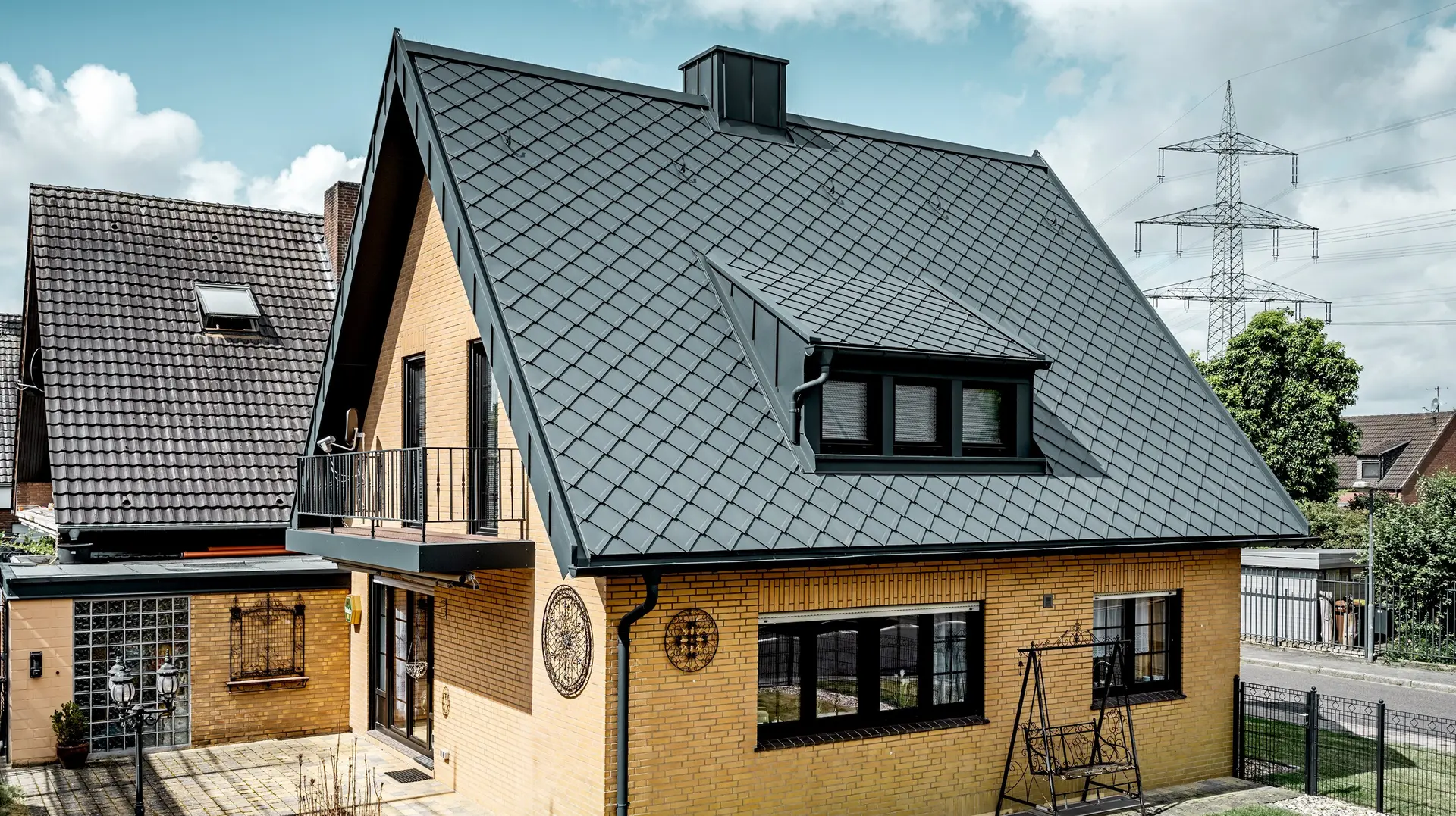 Enfamiljshus i gul klinker efter takrenoveringen med PREFA takromb 29 × 29 i mörkgrått; nybelagt, brant sadeltak med rombmönster, integrerad takkupol och skorsten, terrass och trädgård i förgrunden.