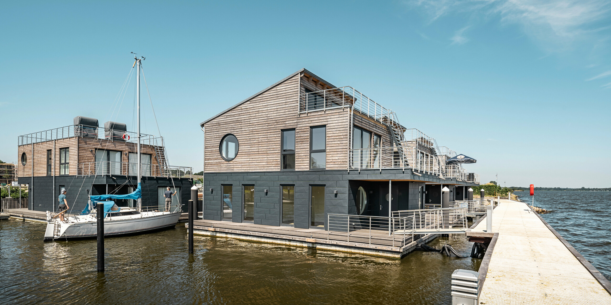 Die harmonische Integration von Architektur und Natur wird in dieser Szene der Wasserhäuser am Pionierhafen Schleswig, mit Blick auf die offene Schlei, sichtbar. Angelegt an einem ruhigen Steg, zeigen die Häuser ihre strapazierfähigen PREFA Siding.X Fassaden in P.10 Anthrazit, die zusammen mit warmen Holzakzenten eine langlebige und wartungsarme Wohnlösung am Wasser bieten. Die idyllische Lage bietet Anwohnern direkten Zugang zum Wasser und eine exklusive Aussicht, ergänzt durch die Ästhetik und Funktionalität der PREFA Aluminiumprodukte.