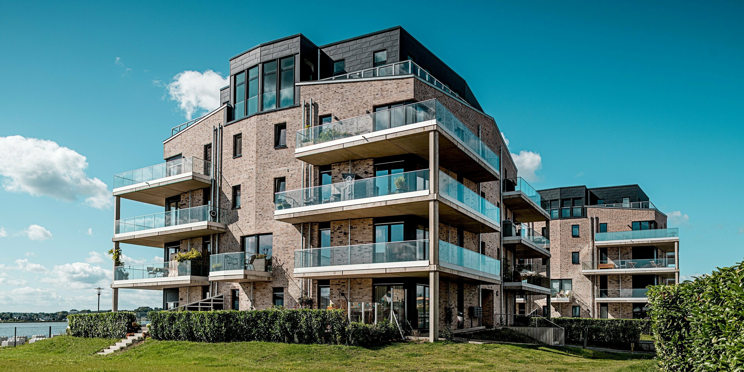 Die Wohnhausanlage „Hafen zur Freiheit“ in Schleswig präsentiert moderne Architektur direkt am Wasser: helle Klinkerfassaden treffen auf dynamisch strukturierte PREFA Siding.X Elemente in P.10 Anthrazit. Besonders auffällig sind die oberen Geschosse mit großzügigen Glasbalustraden und klaren Linien, die im Sonnenlicht elegant kontrastieren. Die widerstandsfähigen Aluminiumfassaden sorgen für langlebigen Schutz vor Küstenwetter und verleihen dem Ensemble einen markanten, hochwertigen Charakter. Die großzügigen Balkone öffnen den Blick auf die maritime Umgebung und unterstreichen das moderne Wohngefühl.