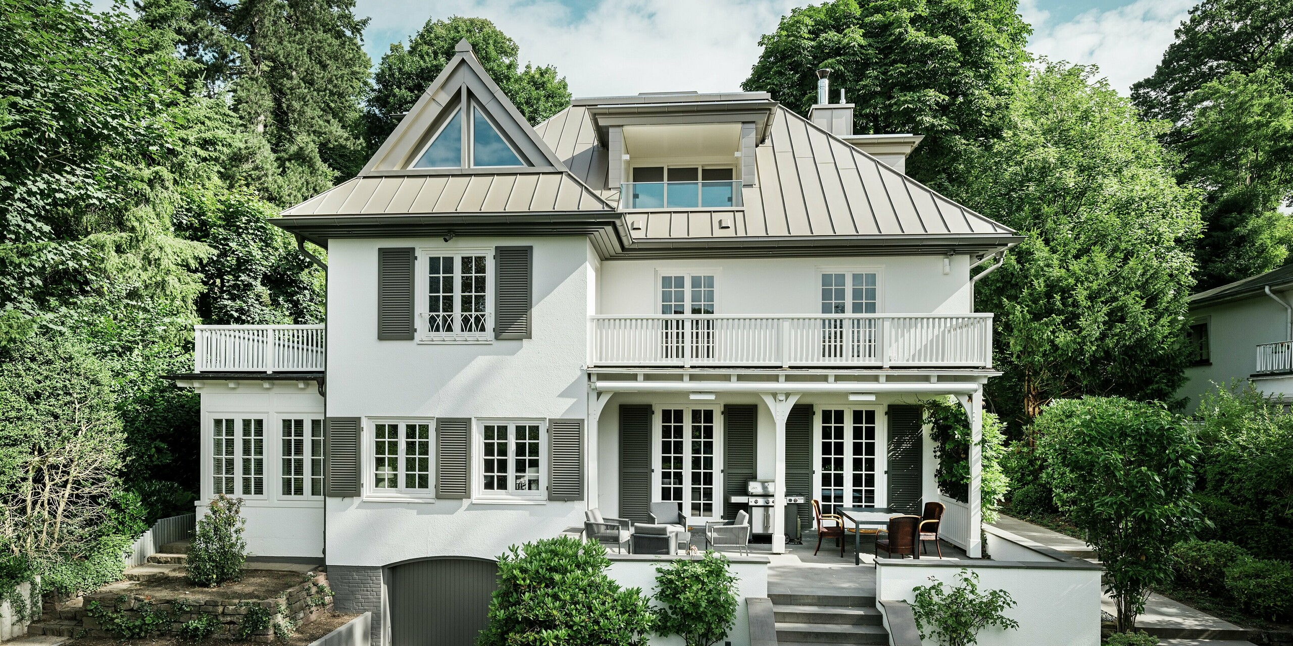 Das Bild zeigt die Villa Blankenese in Hamburg aus einer frontalen Perspektive, eingebettet in üppiges Grün. Das markante Dach aus PREFALZ in Bronze prägt die Architektur und verleiht dem Gebäude eine elegante, zeitlose Ausstrahlung. Die präzise Stehfalztechnik sorgt für eine klare Linienführung und höchste Witterungsbeständigkeit. Ergänzend dazu sind an den Gauben und Fassadenteilen bronzefarbene PREFA Sidings eingesetzt, die das harmonische Zusammenspiel von Dach und Fassade unterstreichen.