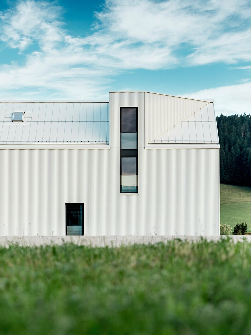 Das Bild zeigt das Einfamilienhaus in Freistadt aus einer Seitenansicht, die die markante, minimalistische Architektur des Gebäudes hervorhebt. Die Fassade aus reinweißem PREFALZ-Aluminium und das steile Satteldach schaffen eine klare, moderne Silhouette, die sich harmonisch in die grüne Umgebung einfügt. Die großen Fensterflächen und das elegante Gaubenfenster betonen das funktionale Design, während das hochwertige Aluminiummaterial sowohl ästhetische Beständigkeit als auch Witterungsresistenz gewährleistet. Das Gebäude strahlt durch seine einfache Form und die Verwendung langlebiger Materialien eine zeitlose Eleganz aus und stellt eine perfekte Kombination aus Architektur und Naturverbundenheit dar.