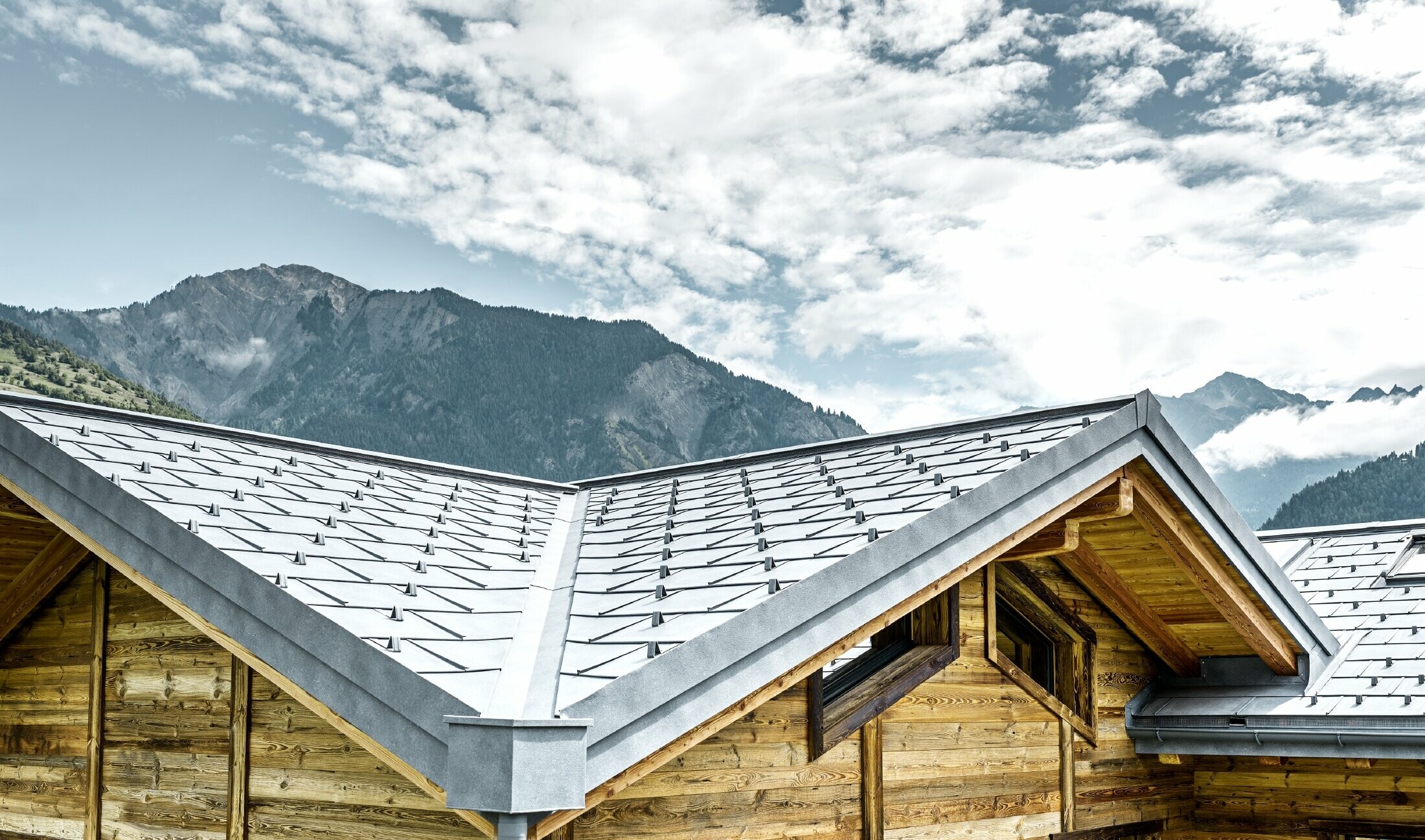 Tak på en rustik stuga i Schweiz med träfasad och ett aluminiumtak från PREFA. Här användes takplattan R.16 i stengrått Man ser det slingrande, vinklade taket och terasstaken.