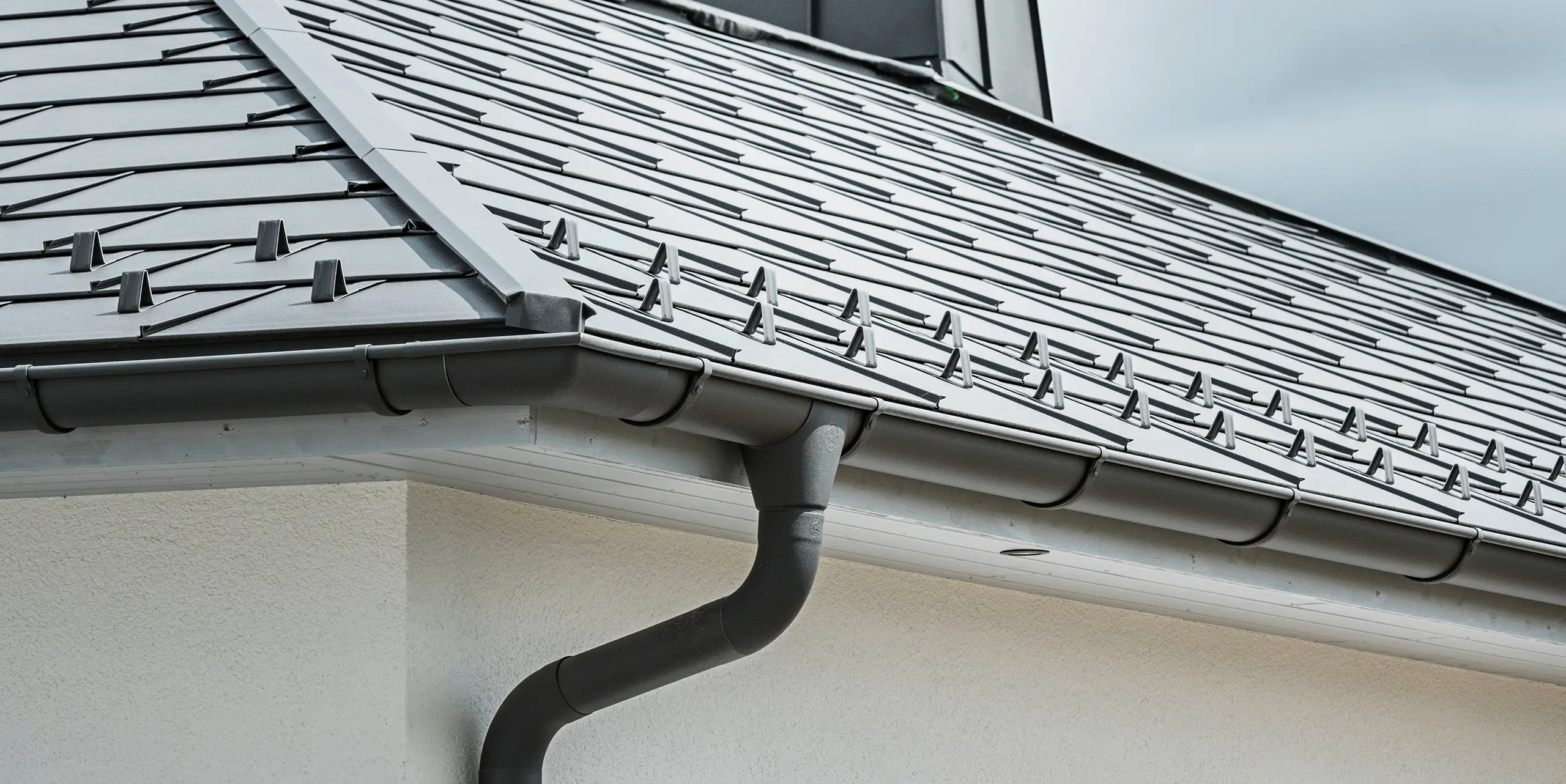Närbild av takfoten på en villa, utrustad med det effektiva PREFA takavvattningssystemet i P.10 mörkgrå. Bilden visar den detaljerade installationen av PREFA Takplatta R.16 och under den den sömlöst integrerade hängrännan samt avloppsröret, som tillsammans erbjuder en pålitlig och estetiskt tilltalande lösning för avledning av regnvatten. De robusta aluminiumprodukterna och den noggranna monteringen understryker kvaliteten och hållbarheten hos PREFA:s kompletta system.