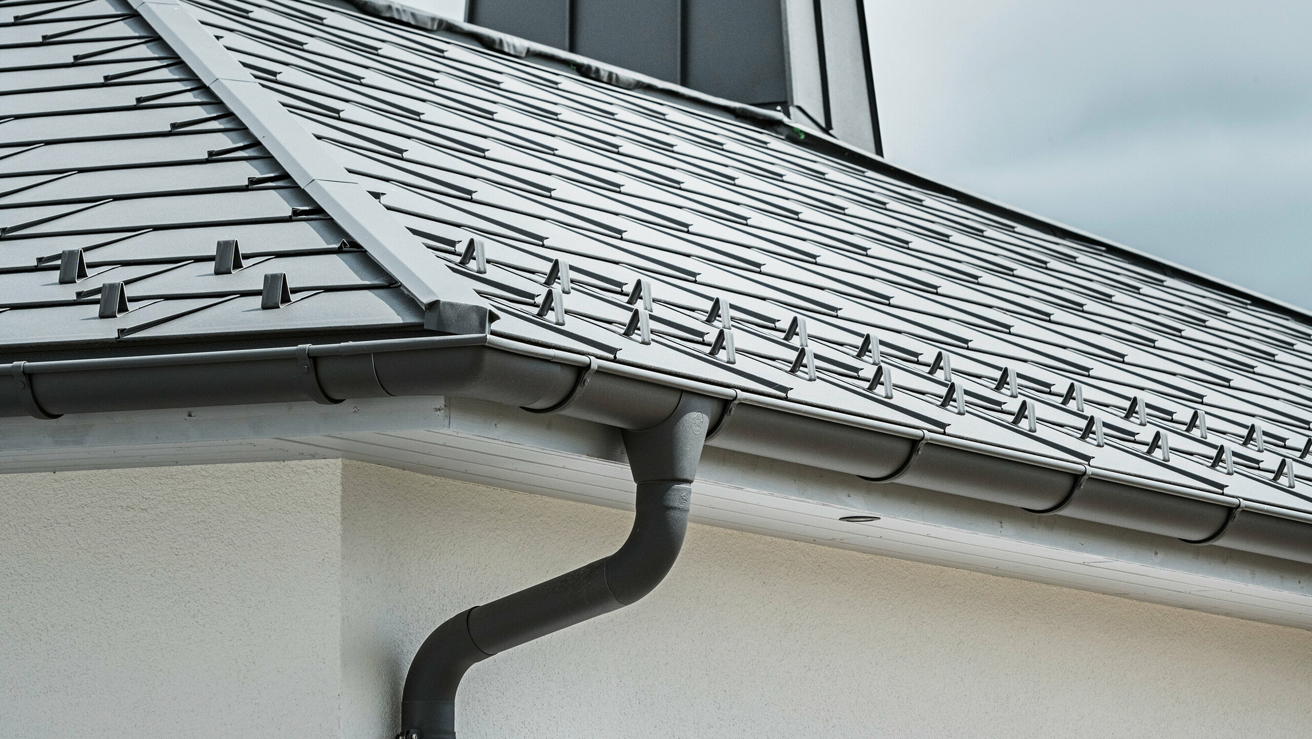 Närbild av takfoten på en villa, utrustad med det effektiva PREFA takavvattningssystemet i P.10 mörkgrå. Bilden visar den detaljerade installationen av PREFA Takplatta R.16 och under den den sömlöst integrerade hängrännan samt avloppsröret, som tillsammans erbjuder en pålitlig och estetiskt tilltalande lösning för avledning av regnvatten. De robusta aluminiumprodukterna och den noggranna monteringen understryker kvaliteten och hållbarheten hos PREFA:s kompletta system.