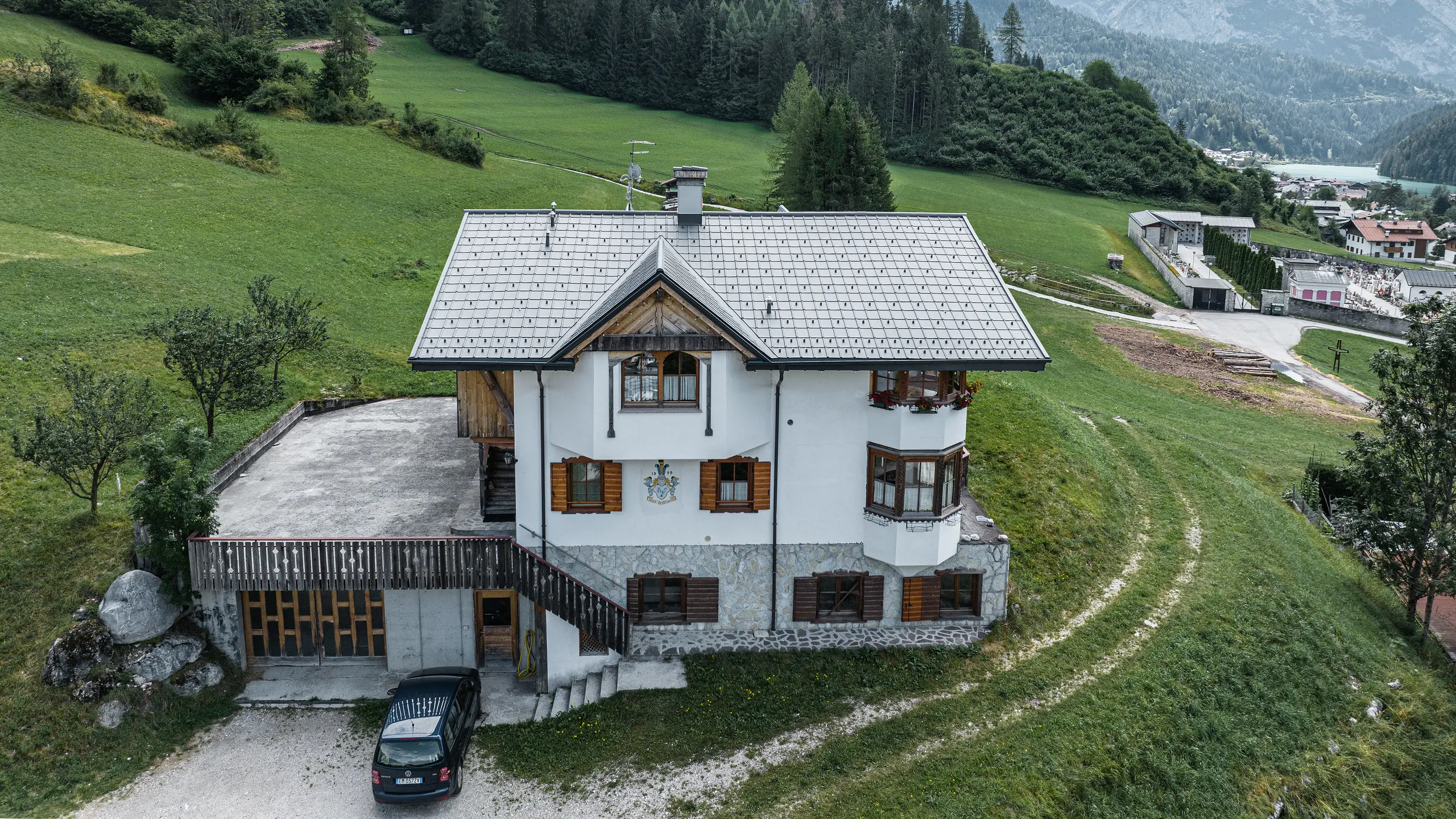 Fågelperspektiv på entrén till den traditionella bergsstugan i Auronzo di Cadore efter takrenoveringen med PREFA aluminiumplattor i ljusgrå P.10. Byggnaden med sin karaktäristiska alpina charm och sitt moderna plåttak smälter harmoniskt in i det grönskande landskapet och det imponerande bergspanoramat i bakgrunden, vilket visar på en lyckad kombination av funktionalitet och regional arkitektur.