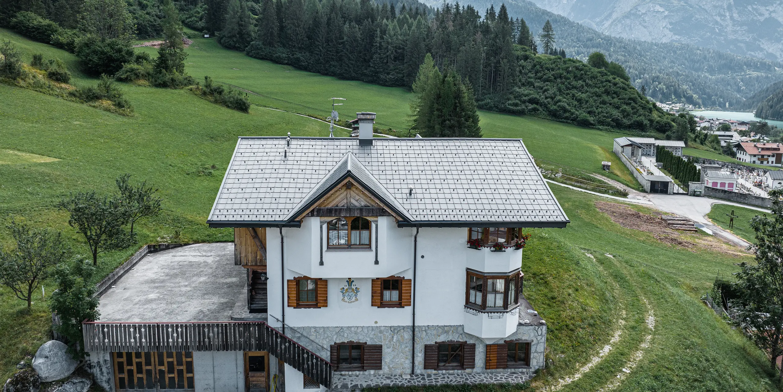 Fågelperspektiv på entrén till den traditionella bergsstugan i Auronzo di Cadore efter takrenoveringen med PREFA aluminiumplattor i ljusgrå P.10. Byggnaden med sin karaktäristiska alpina charm och sitt moderna plåttak smälter harmoniskt in i det grönskande landskapet och det imponerande bergspanoramat i bakgrunden, vilket visar på en lyckad kombination av funktionalitet och regional arkitektur.