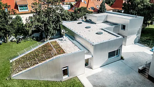 Pri pohľade z vtáčej perspektívy zaujme Envirocentrum Nitra svojou výnimočnou architektúrou a striebristo sa lesknúcou hliníkovou fasádou PREFA z tlačených lisovaných vlnitých profilov v prírodnom hliníkovom vyhotovení. Odolné hliníkové profily s dlhou životnosťou a vysokou odolnosťou voči poveternostným vplyvom spájajú stavebné objekty rôznej výšky do harmonického celku. Zelená šikmá strecha, svetlé ploché strechy a okolité historické budovy zároveň vytvárajú pútavý architektonický kontrast.