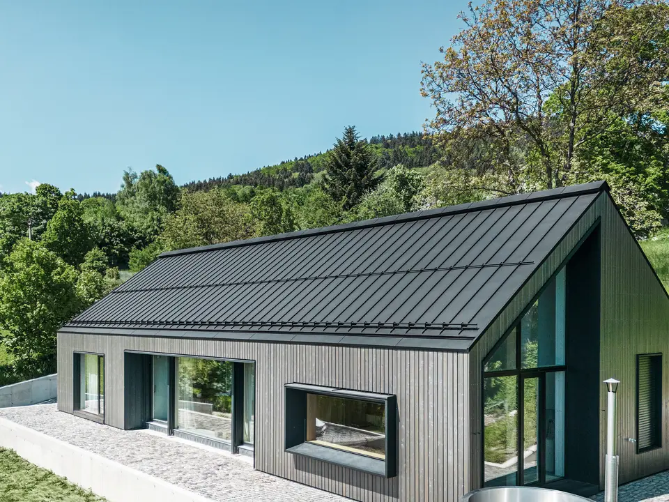 Ein modernes Holzmassivhaus in Kuncice (CZ) wurde mit großen Fensterflächen ausgestattet. Das dunkle Holzmassivhaus verfügt über ein Dachsystem aus PREFALZ in der Farbe P. 10 Schwarz. Im hinteren Außenbereich steht ein Whirlpool mit einer Holzverkleidung.