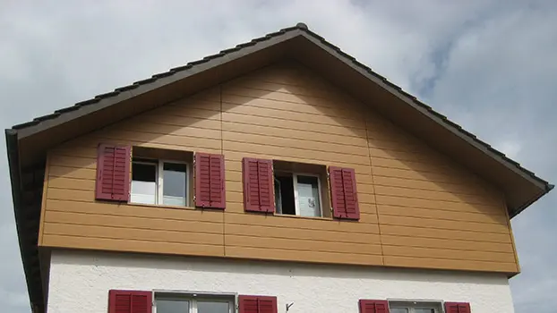 Gavelbeklädnad av ett klassiskt hus med sadeltak. Gaveln är ljust klädd i horisontellt placerade PREFA sidings i trä (ek natur). Fönstren har röda fönsterluckor.