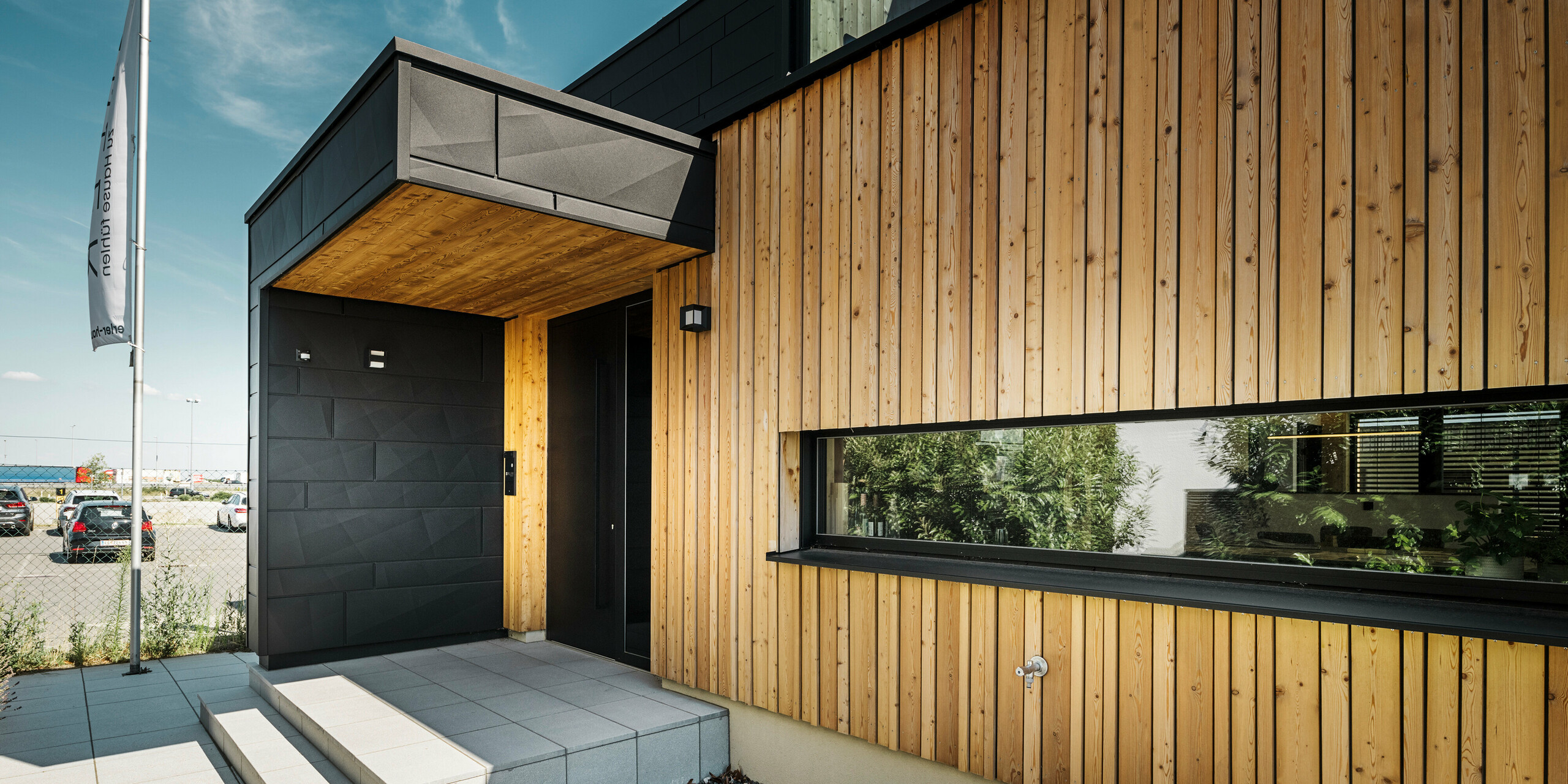Sidoingång till utställningshuset Autark i Blaue Lagunes prefabricerade huscenter i Vösendorf, med en modern fasad av naturligt trä och svarta PREFA Fasadpanel.X-paneler. Bilden visar den eleganta kombinationen av träbeklädnad och aluminium i den högkvalitativa P.10 Black-beläggningen.