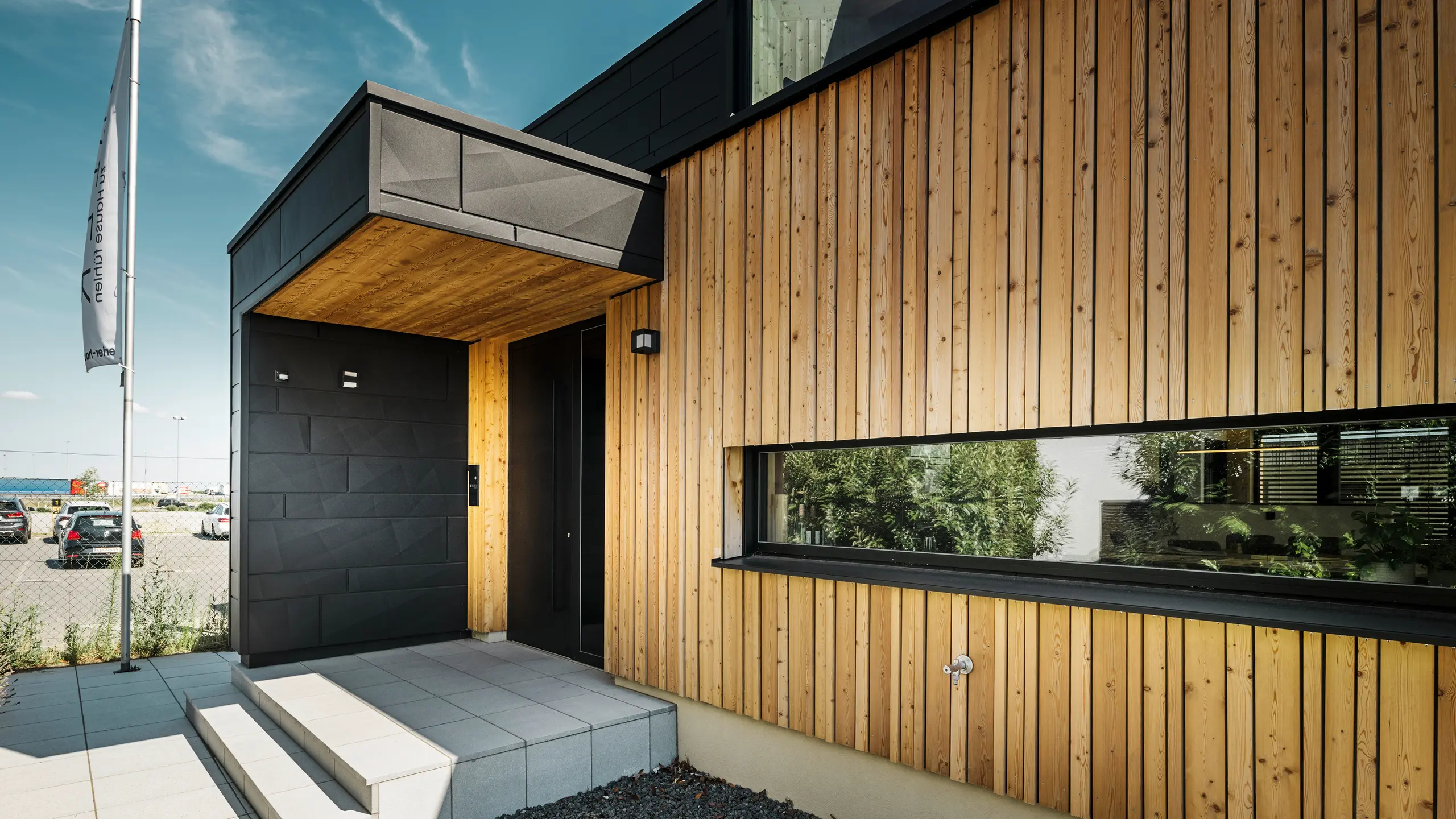Sidoingång till utställningshuset Autark i Blaue Lagunes prefabricerade huscenter i Vösendorf, med en modern fasad av naturligt trä och svarta PREFA Fasadpanel.X-paneler. Bilden visar den eleganta kombinationen av träbeklädnad och aluminium i den högkvalitativa P.10 Black-beläggningen.