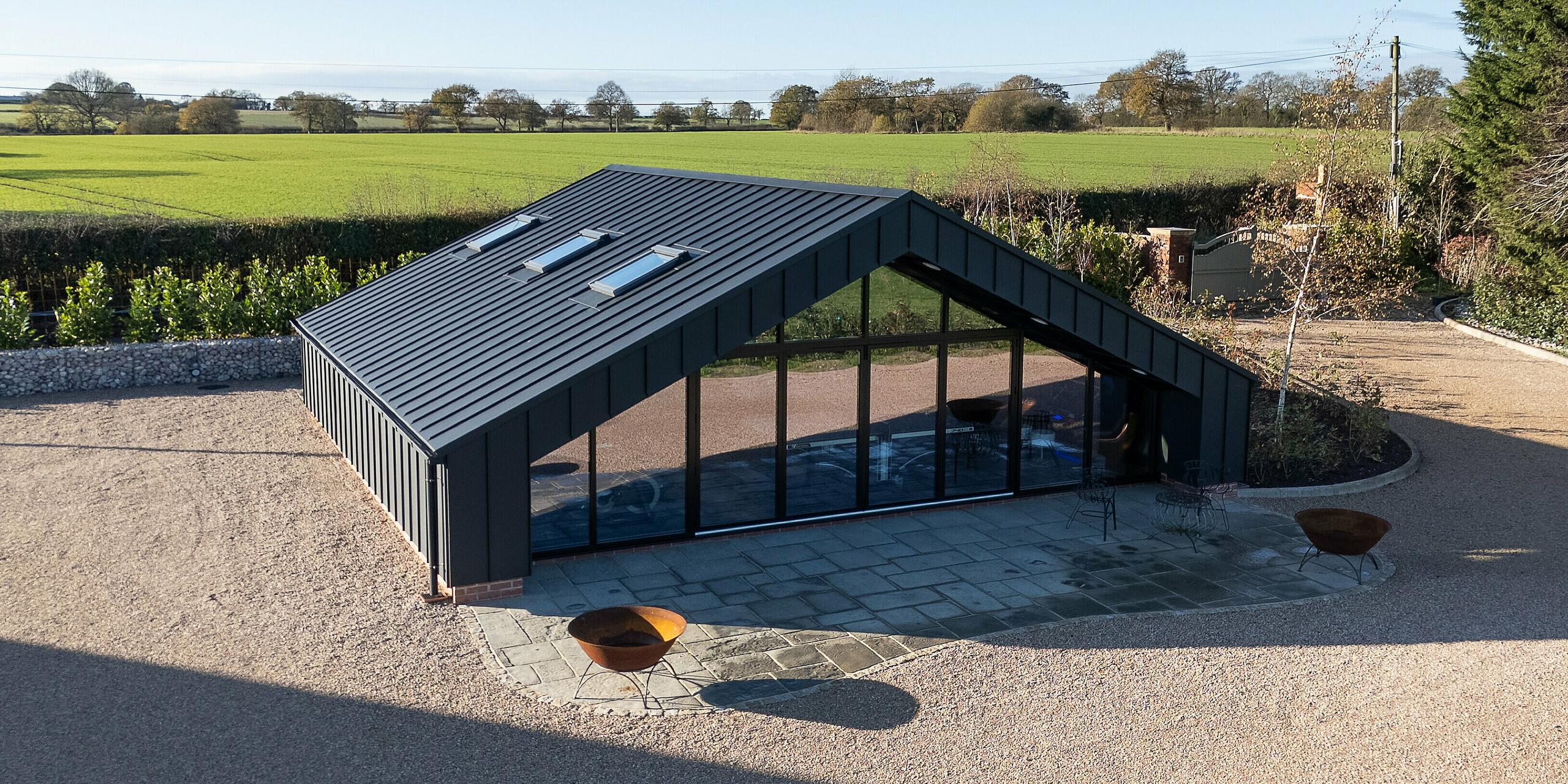 Aerial view of the modern leisure building clad entirely with PREFA aluminum and a PREFALZ standing seam roof in P.10 black. The symmetrical roof shape, expansive glass façade and defined outdoor patio create a striking architectural presence. The durable, weather‑resistant aluminum surface ensures long‑lasting stability and a premium design finish.