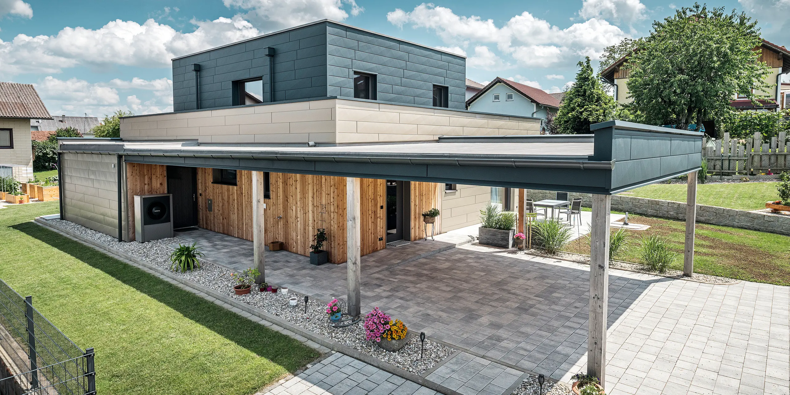 Flygfoto över uppfarten till en modern villa i Wippenham, Österrike, med en tvåfärgad PREFA-aluminiumfasad i brons och antracit. Huset visar den stilfulla integrationen av Fasadpanel.X-fasadpaneler och träbeklädnad. Den täckta carporten, som är sömlöst integrerad i husets arkitektur, är tydligt synlig. Den välskötta trädgården och terrassytan understryker byggnadens moderna och funktionella design.