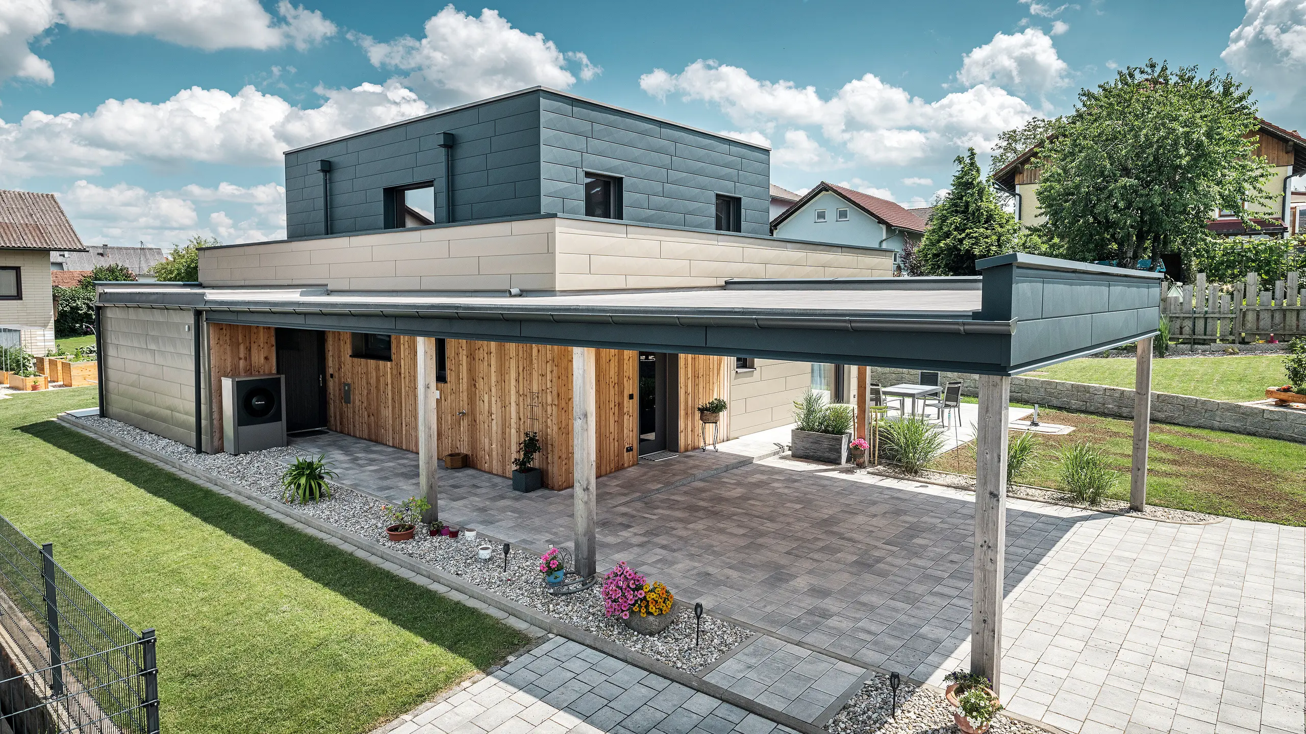 Flygfoto över uppfarten till en modern villa i Wippenham, Österrike, med en tvåfärgad PREFA-aluminiumfasad i brons och antracit. Huset visar den stilfulla integrationen av Fasadpanel.X-fasadpaneler och träbeklädnad. Den täckta carporten, som är sömlöst integrerad i husets arkitektur, är tydligt synlig. Den välskötta trädgården och terrassytan understryker byggnadens moderna och funktionella design.