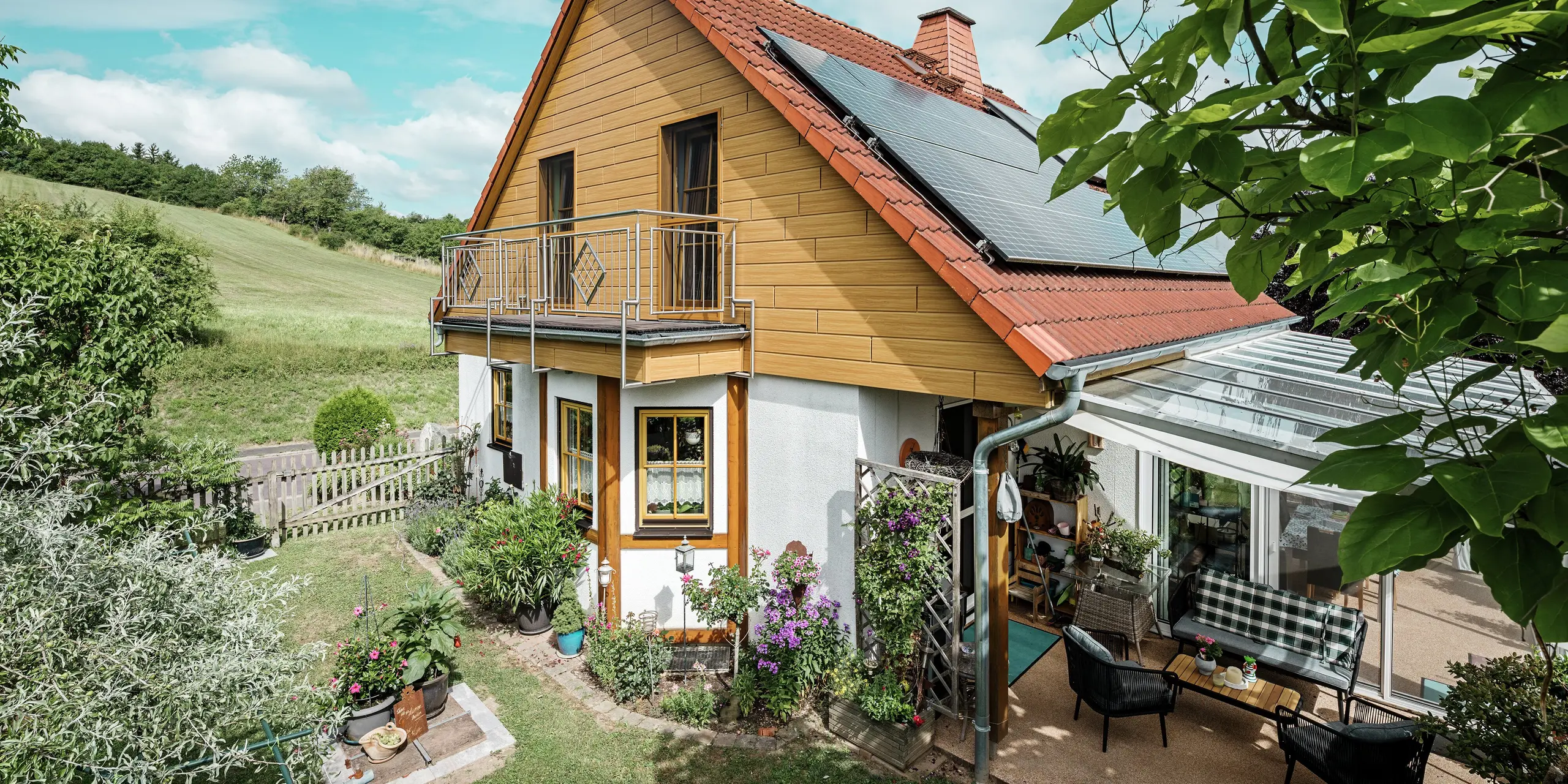 Den här bilden visar ett charmigt enfamiljshus i Blieskastel, som omedelbart fångar blicken med sin nyrenoverade gavel med PREFA Sidings i ek natur. Det varma träutseendet i tålig aluminium harmonierar effektfullt med det röda tegeltaket och det rymliga uterummet. Bland frodig grönska och omsorgsfullt anlagda blomsterrabatter skapas en idyllisk och livfull miljö. Den robusta fasadrenoveringen med PREFA ger inte bara huset ett naturligt utseende, utan imponerar också med sin väderbeständighet, färgtrogenhet och långa, underhållsfria livslängd.