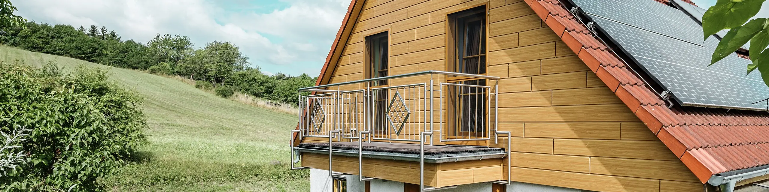 Den här bilden visar ett charmigt enfamiljshus i Blieskastel, som omedelbart fångar blicken med sin nyrenoverade gavel med PREFA Sidings i ek natur. Det varma träutseendet i tålig aluminium harmonierar effektfullt med det röda tegeltaket och det rymliga uterummet. Bland frodig grönska och omsorgsfullt anlagda blomsterrabatter skapas en idyllisk och livfull miljö. Den robusta fasadrenoveringen med PREFA ger inte bara huset ett naturligt utseende, utan imponerar också med sin väderbeständighet, färgtrogenhet och långa, underhållsfria livslängd.