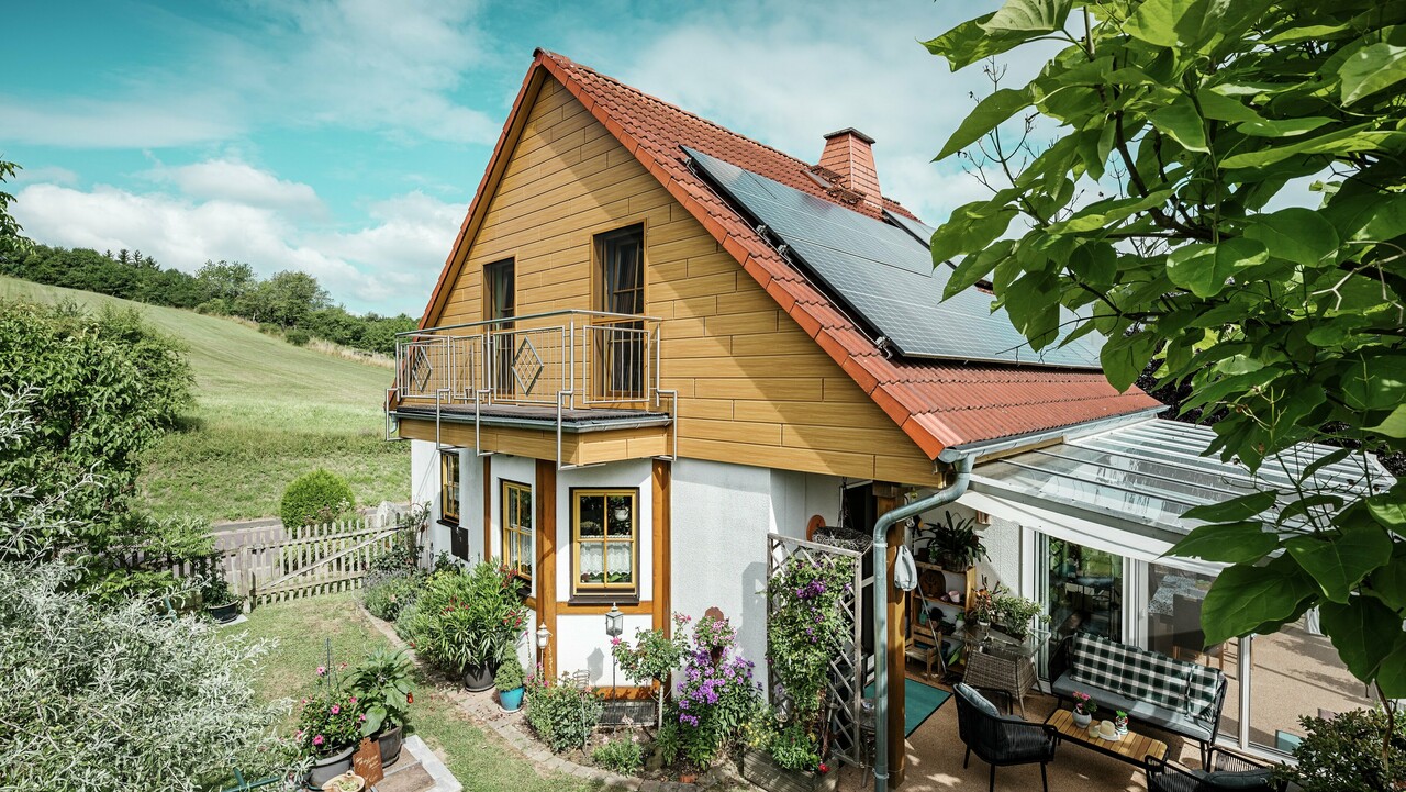 Den här bilden visar ett charmigt enfamiljshus i Blieskastel, som omedelbart fångar blicken med sin nyrenoverade gavel med PREFA Sidings i ek natur. Det varma träutseendet i tålig aluminium harmonierar effektfullt med det röda tegeltaket och det rymliga uterummet. Bland frodig grönska och omsorgsfullt anlagda blomsterrabatter skapas en idyllisk och livfull miljö. Den robusta fasadrenoveringen med PREFA ger inte bara huset ett naturligt utseende, utan imponerar också med sin väderbeständighet, färgtrogenhet och långa, underhållsfria livslängd.