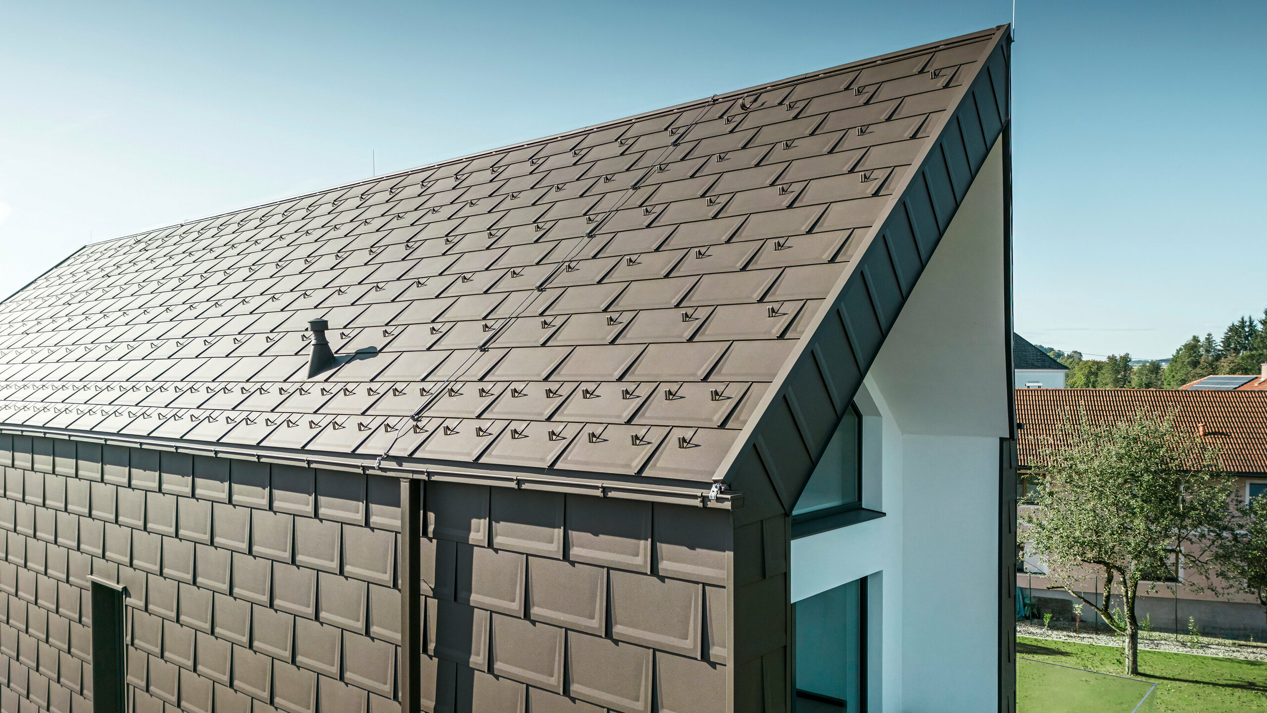 En perspektivvy av tak och fasad på en modern villa i Neukirchen, Österrike. PREFA Takplatta R.16 i brun nyans P.10 erbjuder en estetisk och funktionell takbeläggning som passar perfekt in i husets arkitektoniska utformning. Den skarpa taknocken och den exakta placeringen av takplattorna visar PREFAs kvalitet. De små aluminiumprodukterna smälter perfekt in i den vitputsade fasaden och det omgivande gröna landskapet.