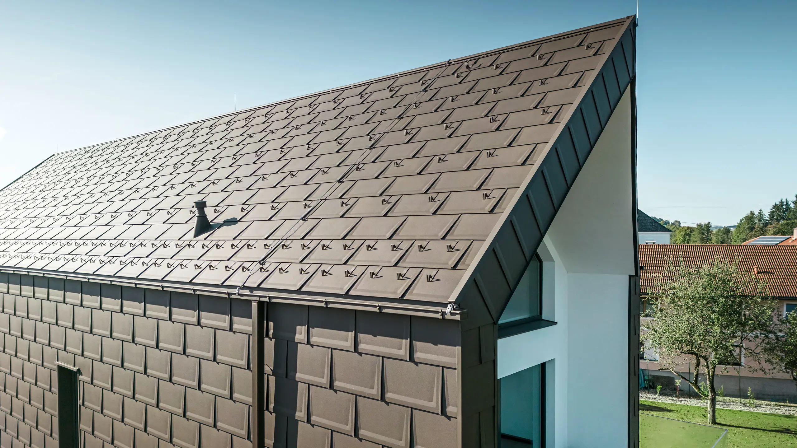 En perspektivvy av tak och fasad på en modern villa i Neukirchen, Österrike. PREFA Takplatta R.16 i brun nyans P.10 erbjuder en estetisk och funktionell takbeläggning som passar perfekt in i husets arkitektoniska utformning. Den skarpa taknocken och den exakta placeringen av takplattorna visar PREFAs kvalitet. De små aluminiumprodukterna smälter perfekt in i den vitputsade fasaden och det omgivande gröna landskapet.