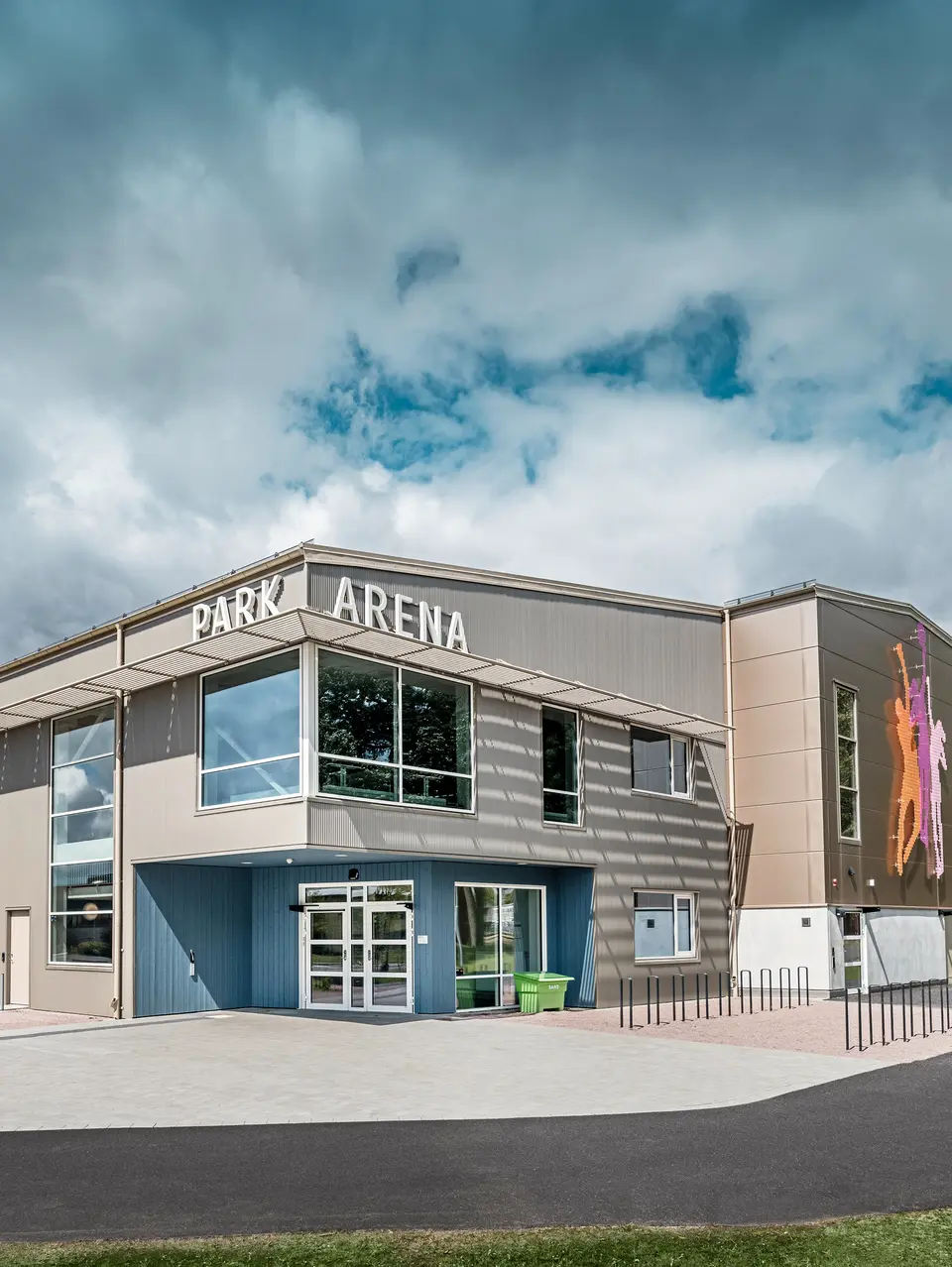 Frontansicht der Park Arena in Värnamo, Schweden, mit moderner PREFA Fassade aus Zackenprofilen in Bronze. Die Aluminiumfassade bietet eine langlebige und wetterbeständige Lösung, die durch markante Licht- und Schatteneffekte hervorsticht. Ein künstlerisch gestaltetes Wandbild auf der rechten Seite zeigt farbenfrohe sportliche Figuren, die den dynamischen Charakter der Sporthalle betonen. Große Fensterflächen lassen viel natürliches Licht in das Gebäude und verstärken das moderne Design der Arena.