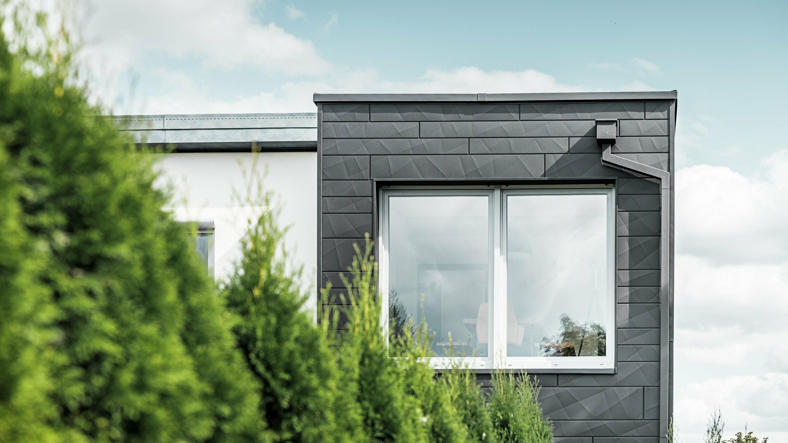 De tydligt strukturerade PREFA Fasadpanel.X fasadpanelerna i färgen P.10 svart pryder ytterväggen på en villa i Teuschnitz och ger den ett unikt utseende. PREFA takavvattning, bestående av fyrkantrör och vattenuppsamlingslåda, smälter sömlöst in i designen. De gröna tuja-träden i förgrunden kontrasterar mot den mörka plåtfasaden.