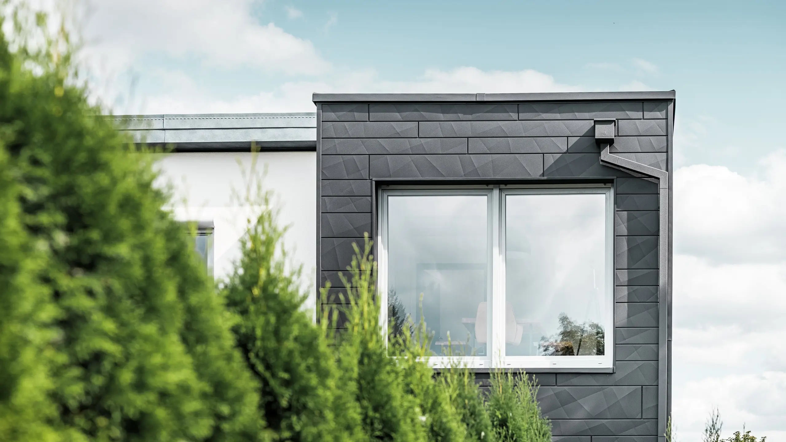 De tydligt strukturerade PREFA Fasadpanel.X fasadpanelerna i färgen P.10 svart pryder ytterväggen på en villa i Teuschnitz och ger den ett unikt utseende. PREFA takavvattning, bestående av fyrkantrör och vattenuppsamlingslåda, smälter sömlöst in i designen. De gröna tuja-träden i förgrunden kontrasterar mot den mörka plåtfasaden.