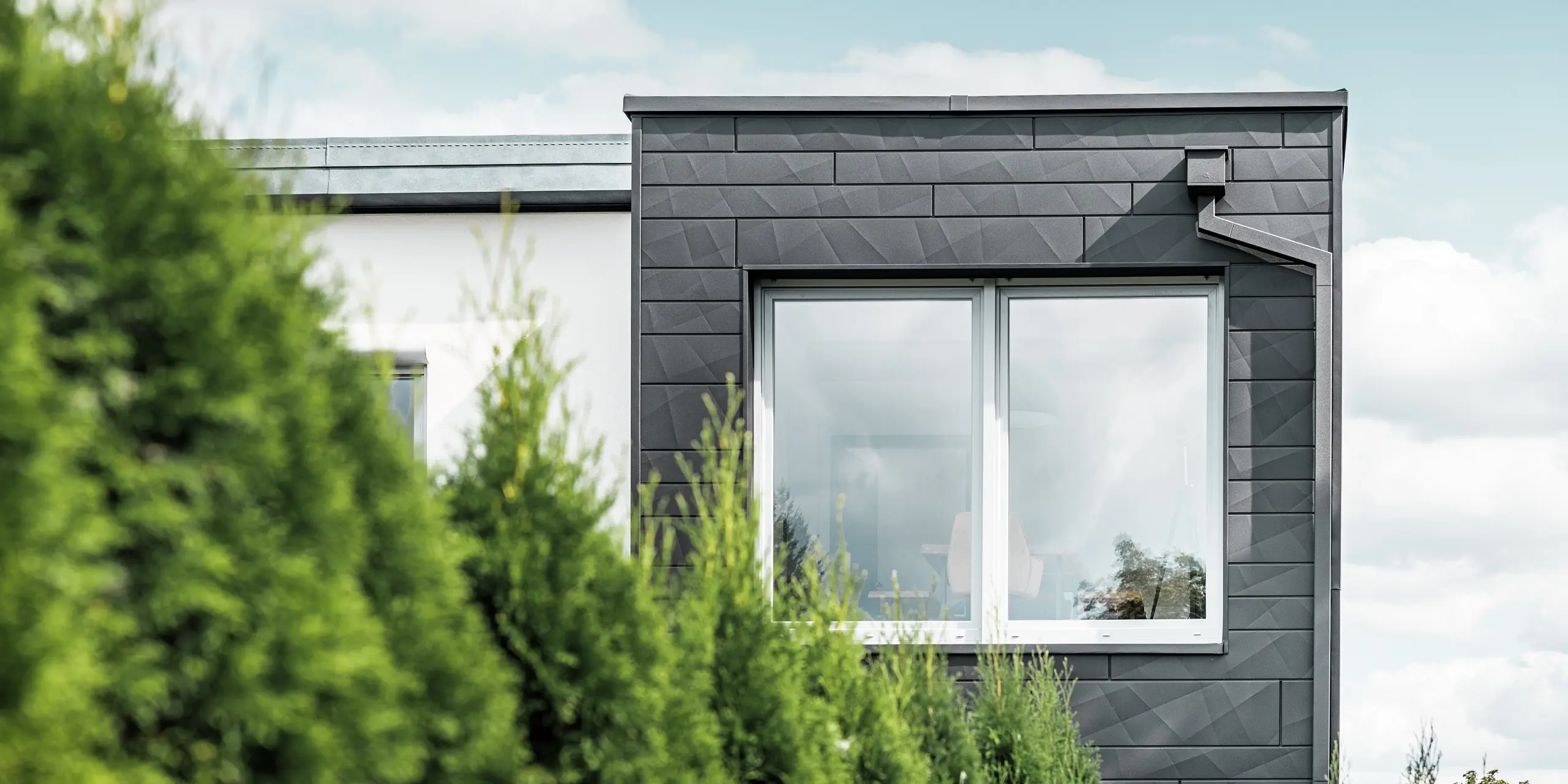 De tydligt strukturerade PREFA Fasadpanel.X fasadpanelerna i färgen P.10 svart pryder ytterväggen på en villa i Teuschnitz och ger den ett unikt utseende. PREFA takavvattning, bestående av fyrkantrör och vattenuppsamlingslåda, smälter sömlöst in i designen. De gröna tuja-träden i förgrunden kontrasterar mot den mörka plåtfasaden.