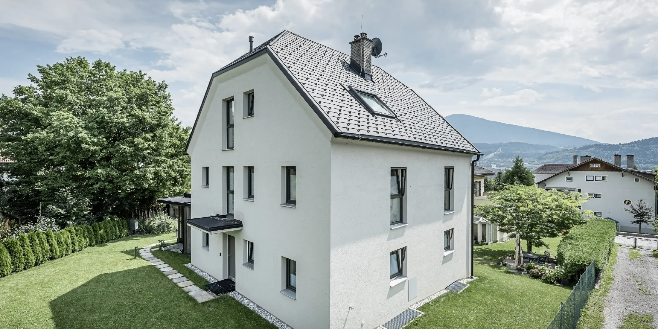 Modernt trevåningshus med ljus putsad fasad och mörkgrå sadeltak av aluminium från PREFA. Byggnaden har flera smala fönster och takavvattning i antracit. Den ligger i en välskött trädgård med gräsmatta, träd och häckar, och i bakgrunden syns ytterligare bostadshus och ett bergigt landskap.